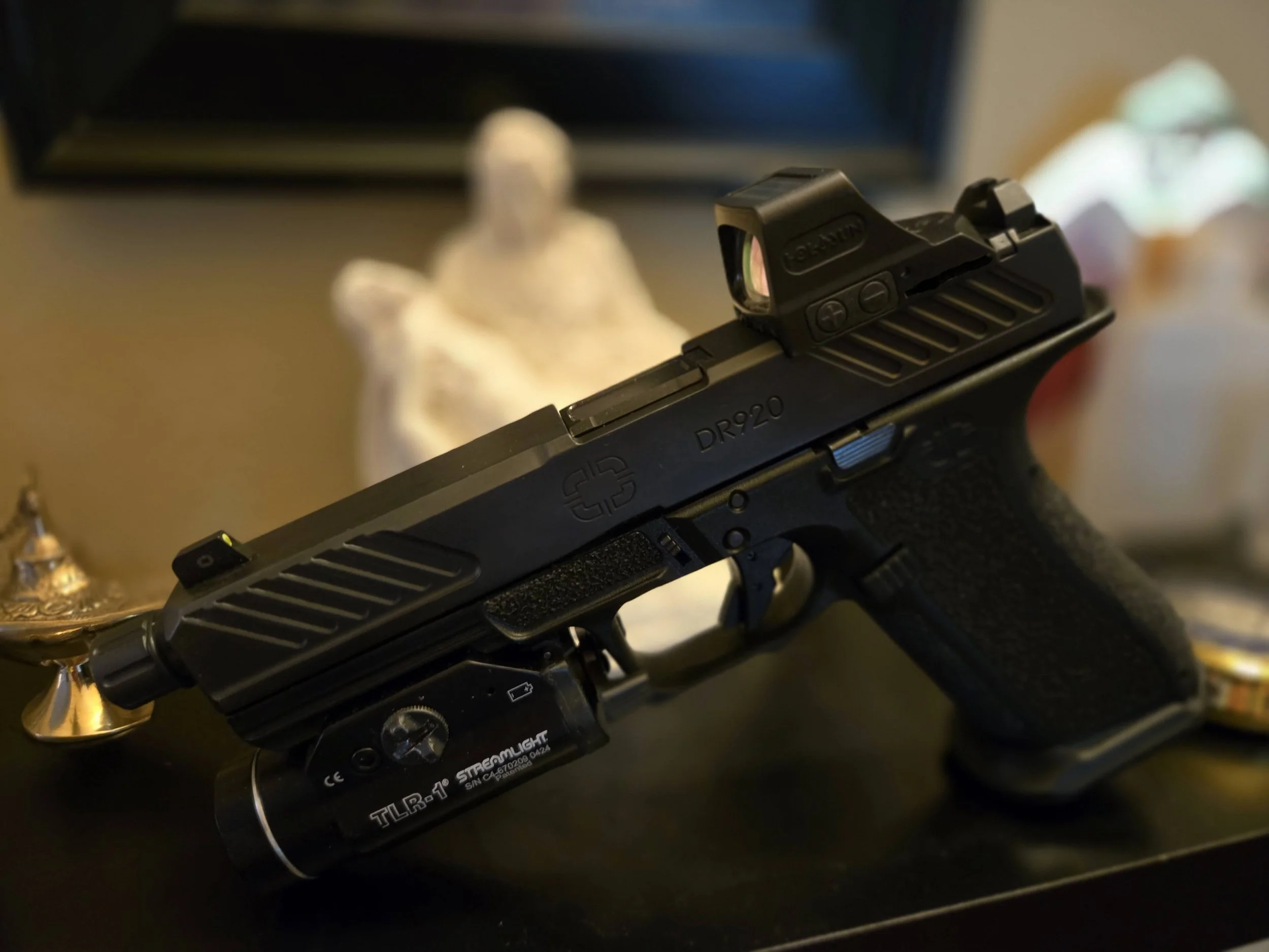 A black handgun with a mounted red dot sight, placed on a surface with decorative items and a blurred background.