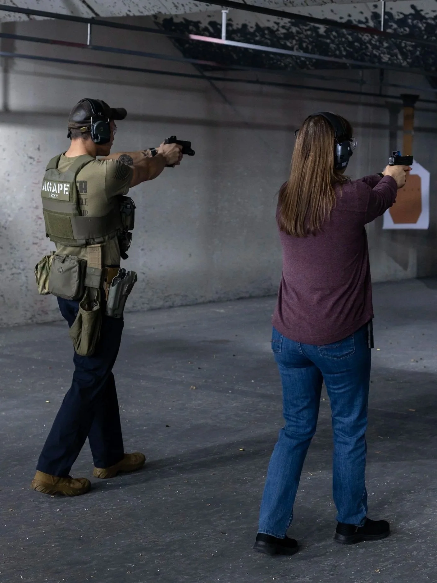 Two individuals at a shooting range aiming pistols at targets, wearing hearing protection and casual clothing.