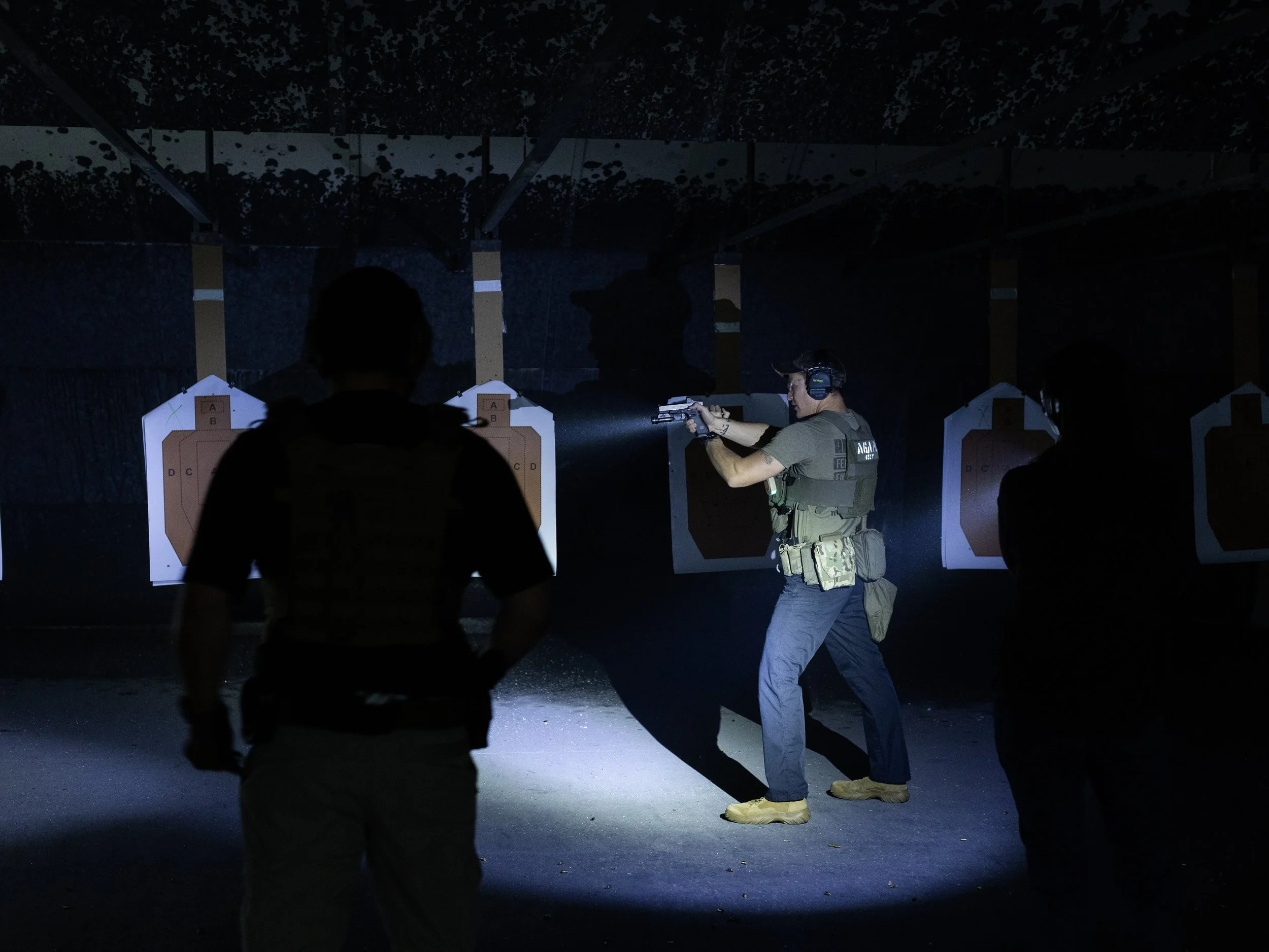A person wearing tactical gear and ear protection shooting a handgun during a training session at an indoor firing range with targets in the background, illuminated by a flashlight.