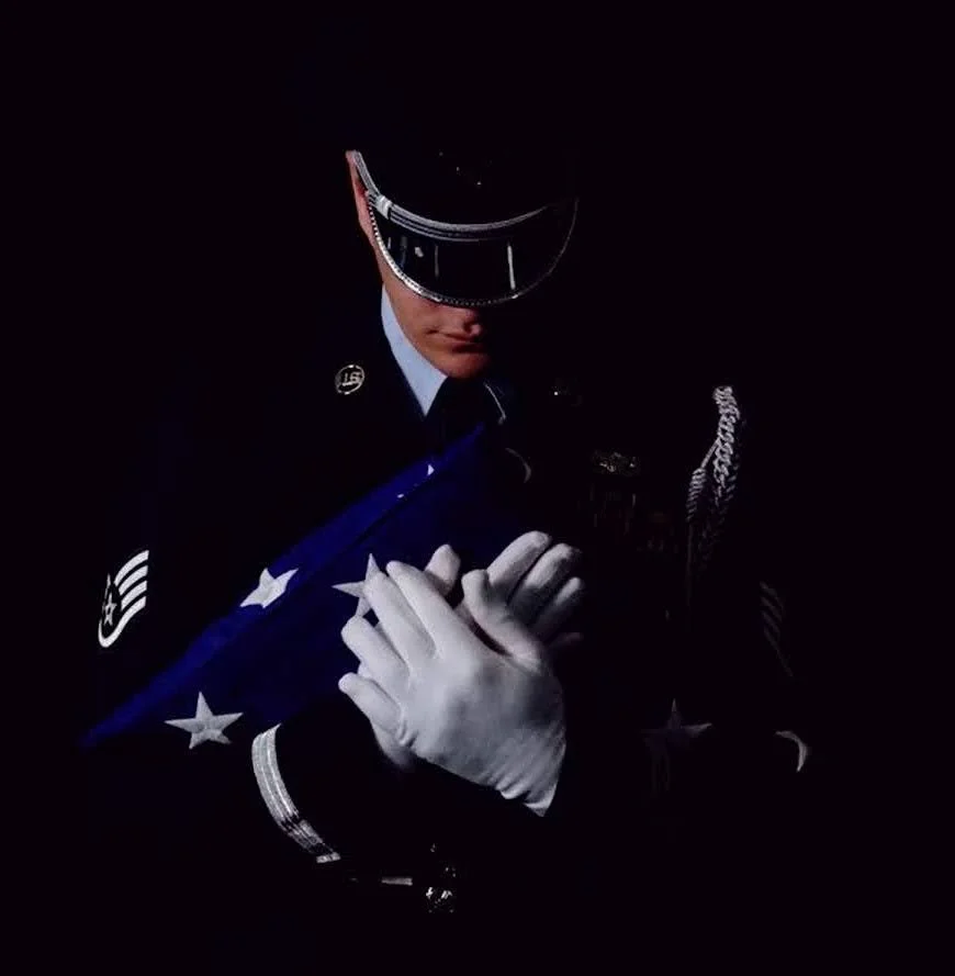 A person in a navy uniform with white gloves, holding a folded American flag against a dark background.