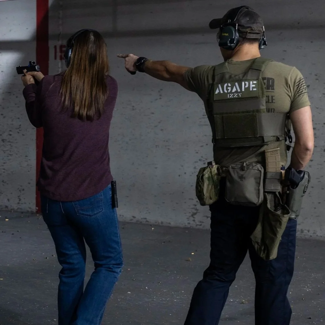 A woman holding a handgun with both hands and a man in a tactical vest and headset issuing commands at her in a shooting range.