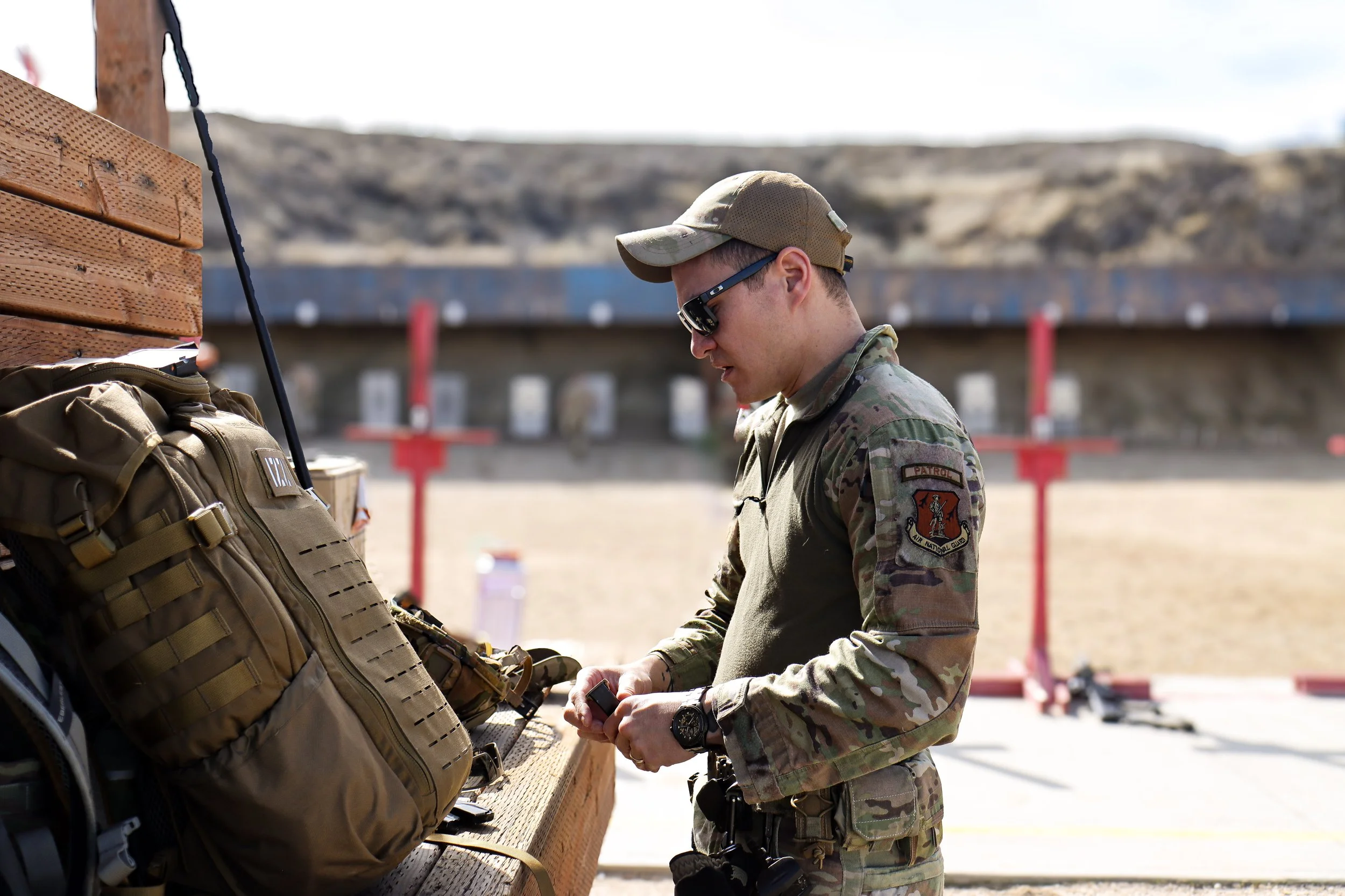 Soldier in camouflage uniform and sunglasses at outdoor shooting range, inspecting equipment on a wooden table with firearms targets in the background.