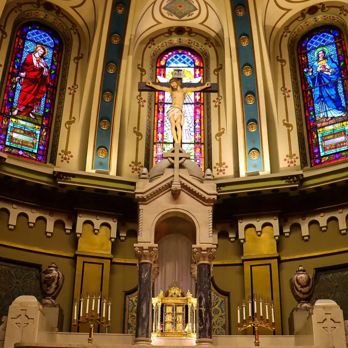 The interior of a church with stained glass windows depicting religious figures, a crucifix with Jesus Christ, and an ornate altar with candles and decorations.