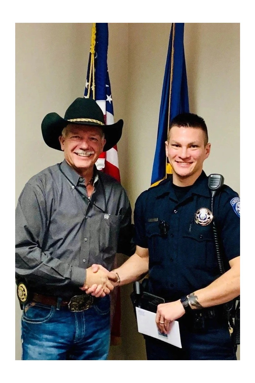 Two men shaking hands in front of American flags, one dressed as a cowboy and the other as a police officer, both smiling.