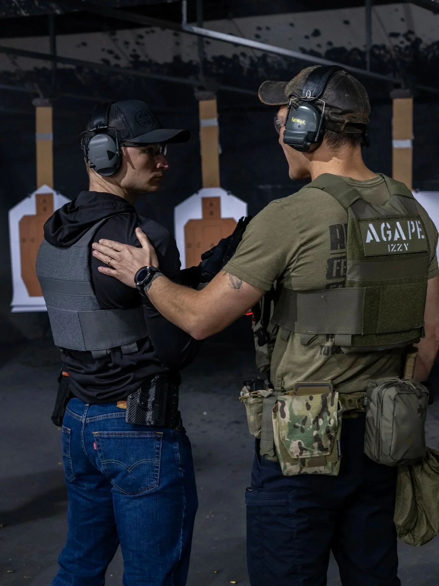 Two men at an indoor shooting range wearing hearing protection and tactical gear, with target silhouettes in the background.