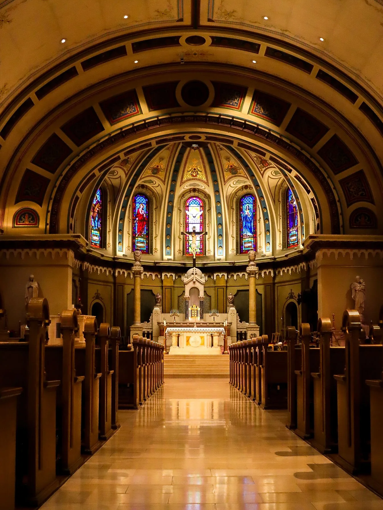 Interior of a church with an altar at the front, stained glass windows with religious images, and a domed ceiling with ornate artwork.