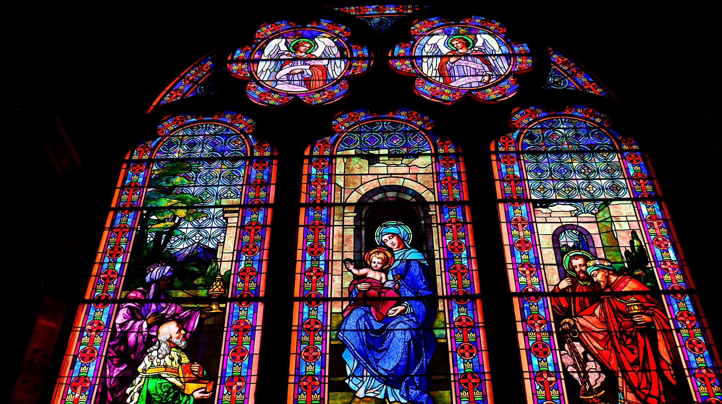 Colorful stained glass church window depicting religious figures, including the Virgin Mary holding baby Jesus, surrounded by angels and saints, with intricate geometric and floral patterns.