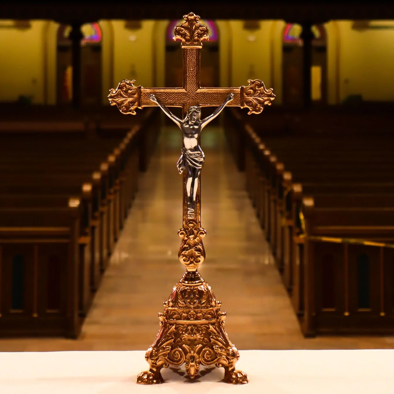 A gold-colored crucifix with a detailed base, set on a white surface inside a church with wooden pews and stained glass windows in the background.