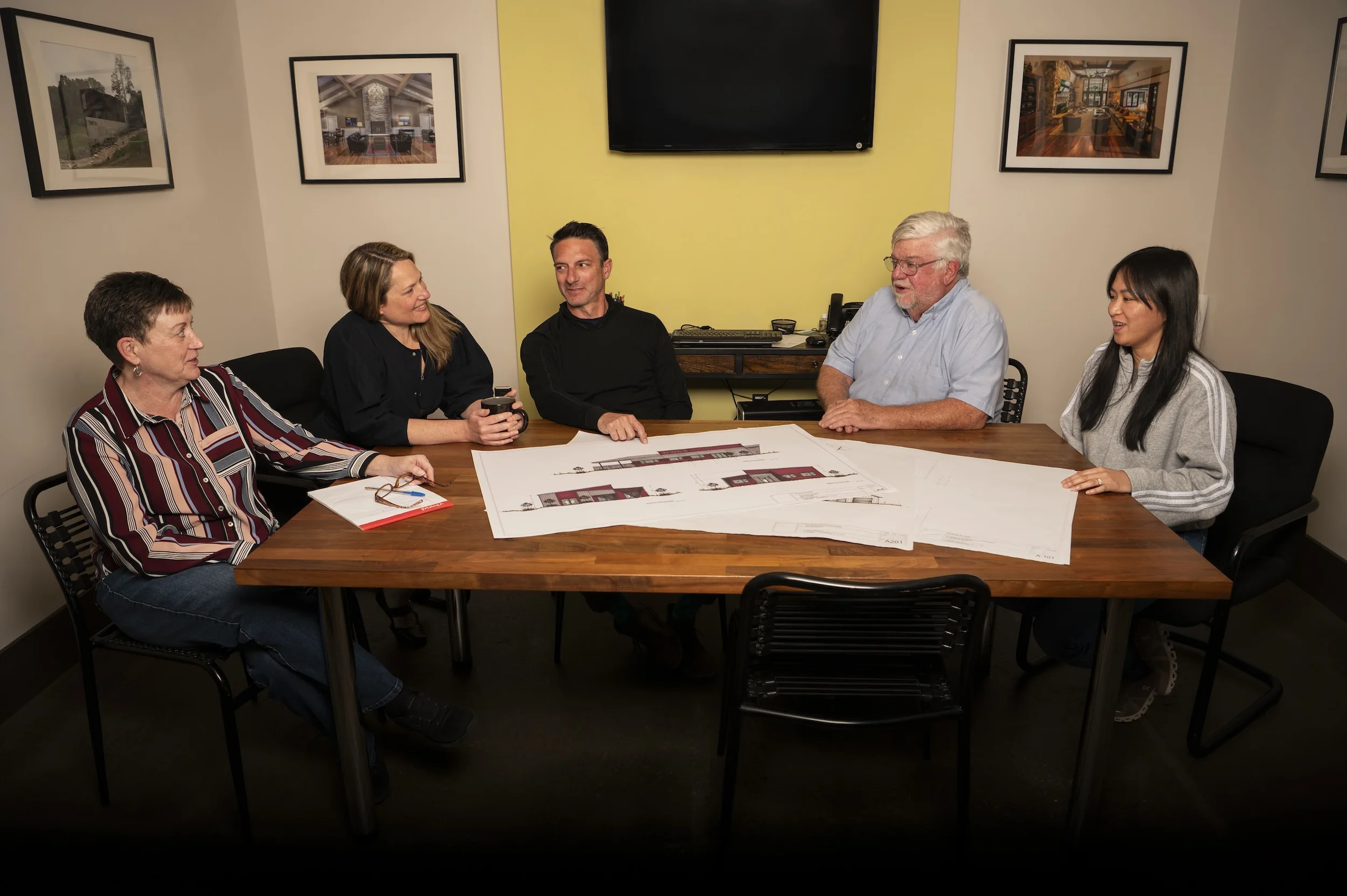 A group of six people sitting around a table in a conference room, discussing architectural plans with blueprints spread out on the table. There are framed photos of interiors and exteriors on the walls, and a large television mounted on the wall behind them.