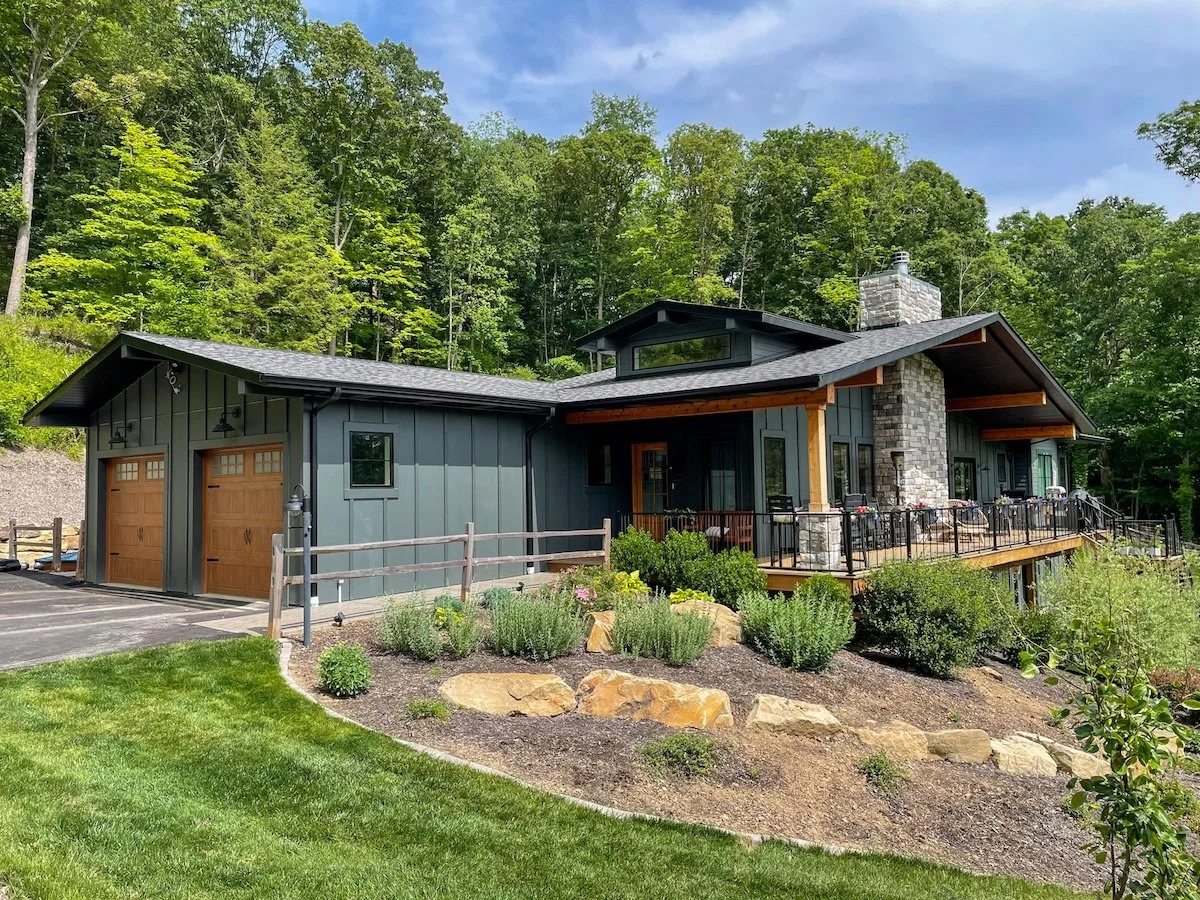 Modern house with a stone chimney, grey and wood exterior, and a large deck overlooking a garden and lush green forest.