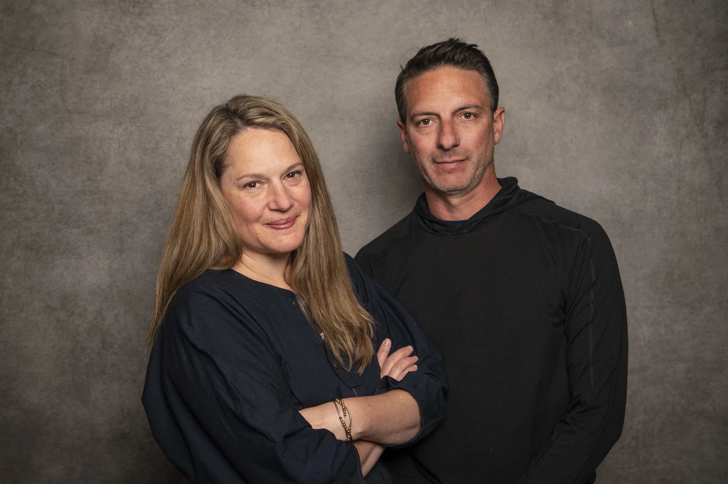 A woman with long red hair and a man with short dark hair standing close against a gray background, both looking at the camera with slight smiles.
