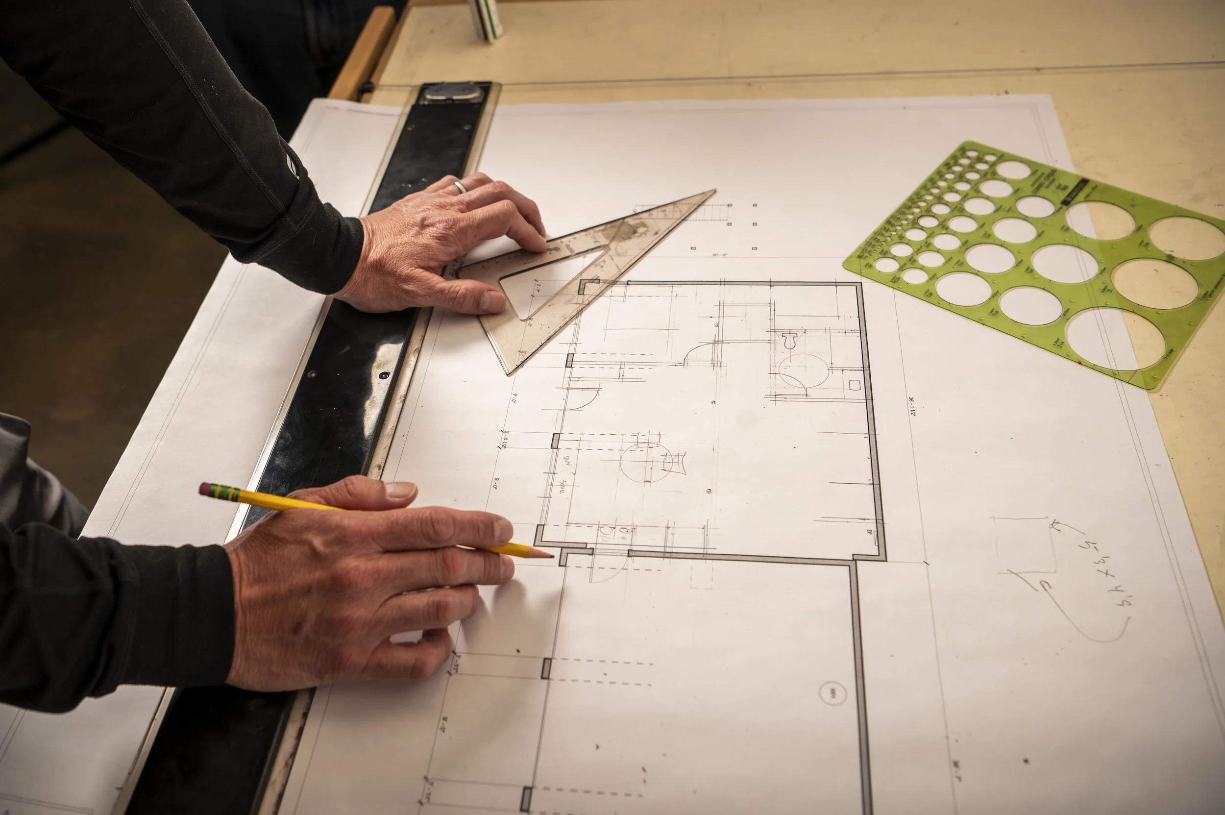A person is working on architectural plans or blueprints, using a triangular ruler and a pencil, with drafting tools such as a circle template and a straightedge on the table.