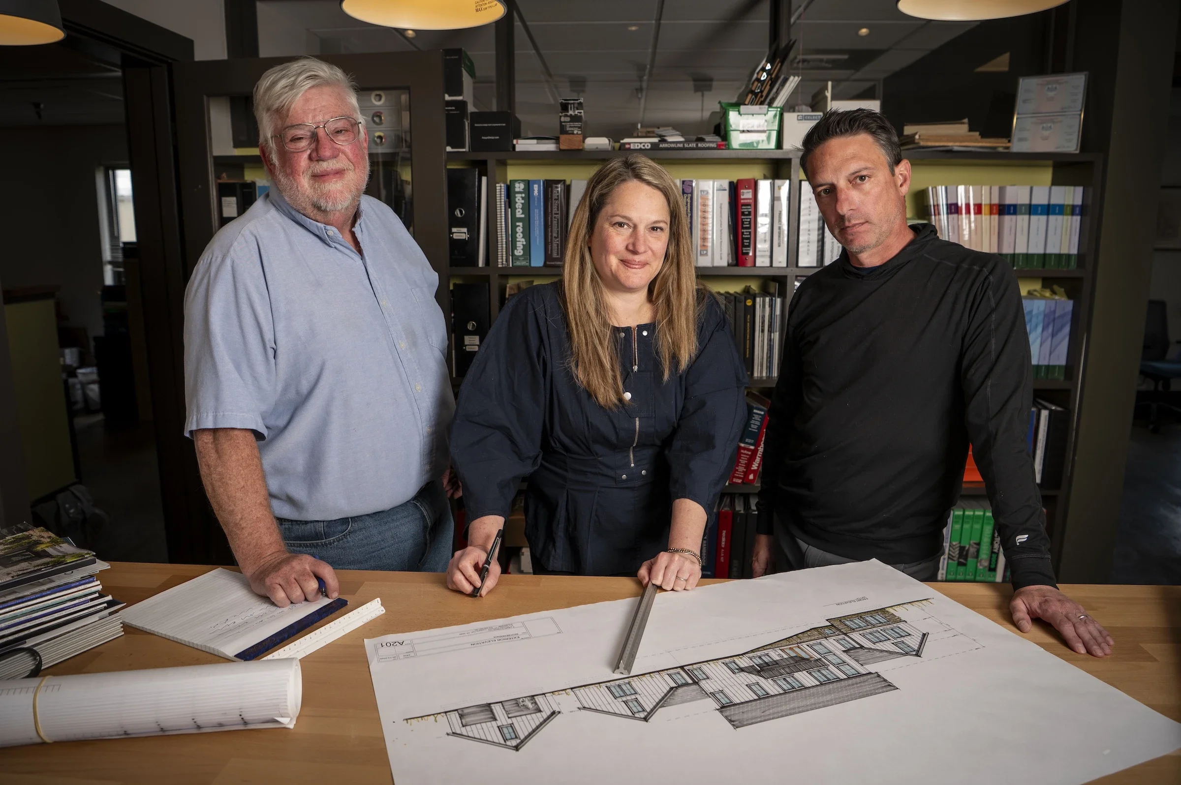 Three professionals stand behind a table reviewing architectural blueprints in an office with bookshelves filled with binders and books.