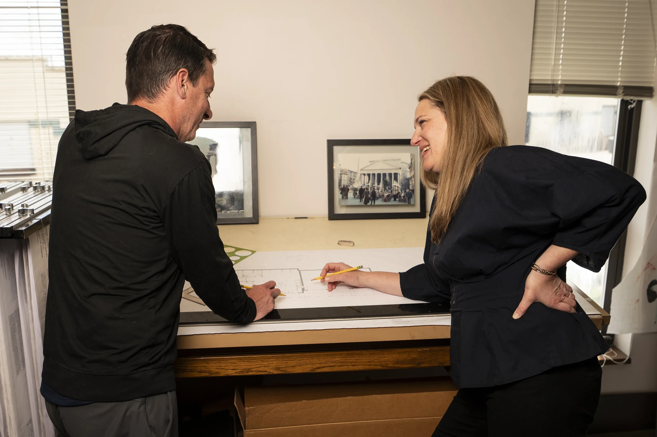 A man and woman are engaged in a discussion over architectural plans, leaning on a table with framed artwork of city scenes in the background.
