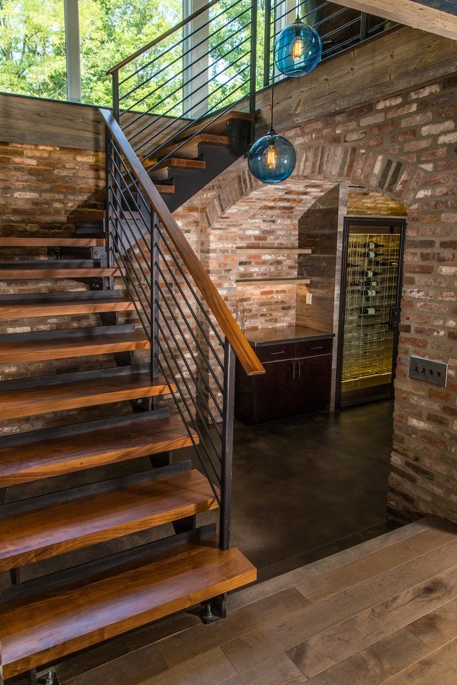 Interior view of a modern home featuring a staircase with wooden steps and black metal railing, exposed brick walls, and two blue pendant lights hanging from the ceiling. There is a wine cooler and a dark wooden cabinet below the staircase.