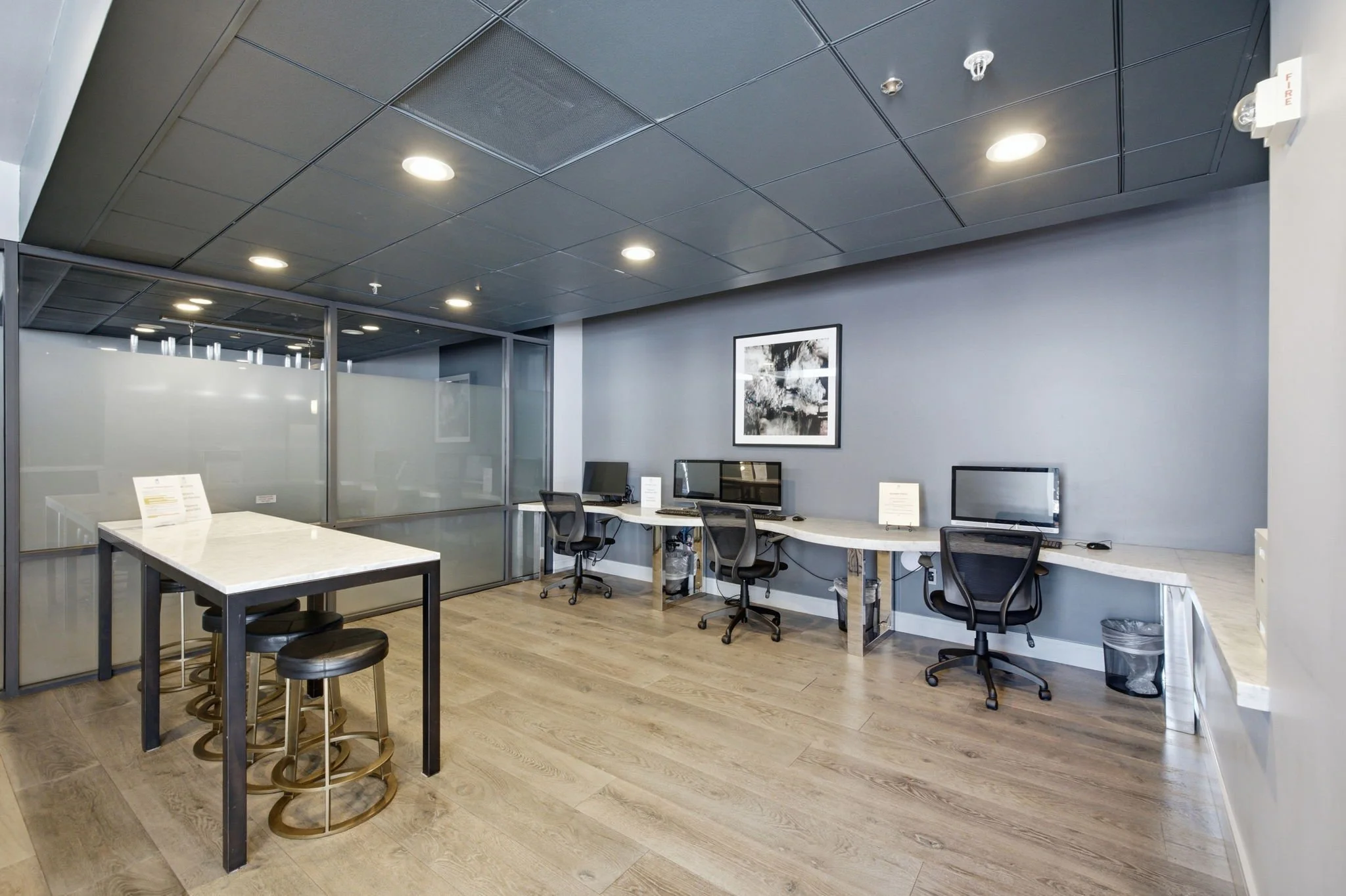 Office workspace with three computer stations along a curved desk, a small high table with three stools, and a black-and-white framed picture on a gray wall. Hardwood flooring and a black suspended ceiling with recessed lighting.