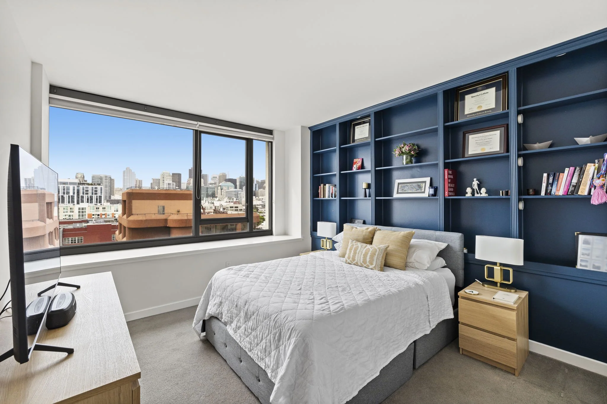 Modern bedroom with large window showing city skyline, blue accent wall with built-in shelves, bed with white quilt and beige pillows, wooden nightstand, and small TV on a wooden dresser.