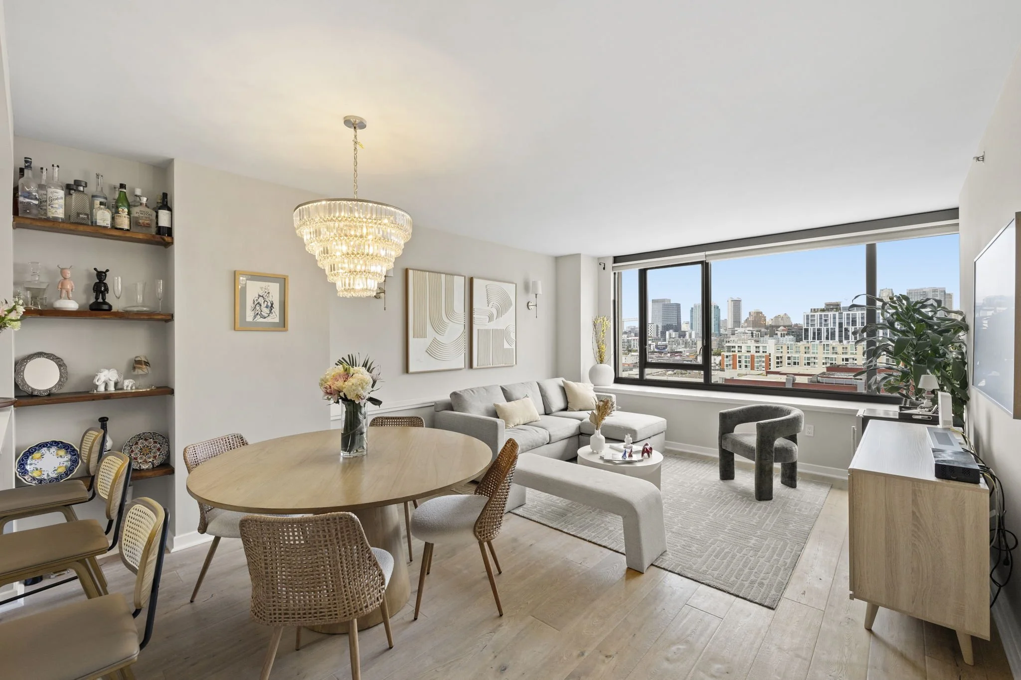 Modern living room with large window overlooking city skyline, featuring a gray sectional sofa, decorative wall art, a round dining table with chairs, and a chandelier.