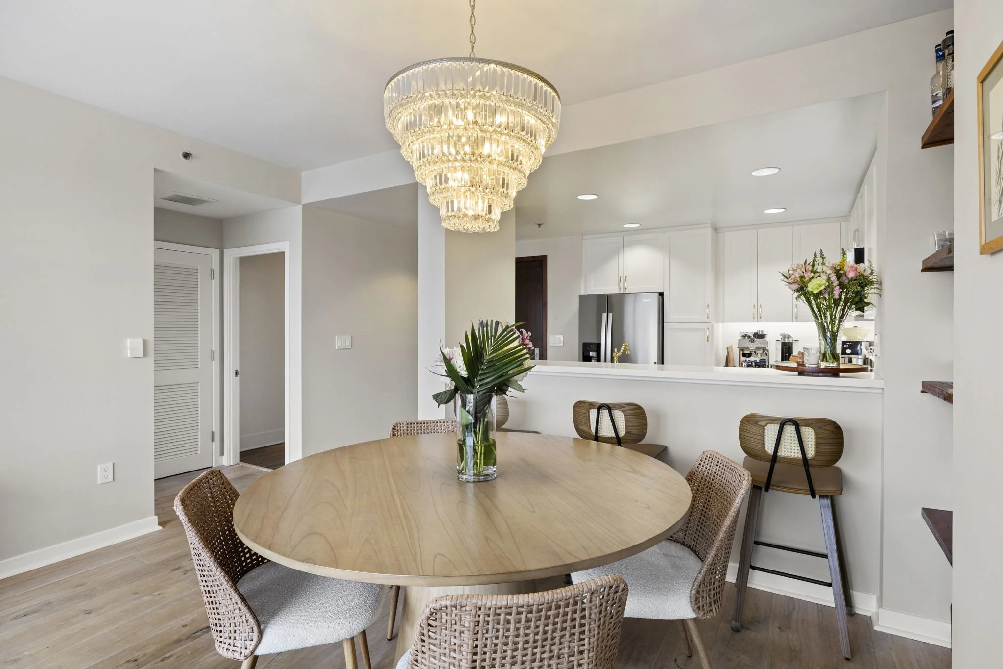 Modern dining and kitchen area with a round wooden table, wicker chairs, a chandelier, and a kitchen with white cabinets and stainless steel appliances, decorated with vases of flowers.