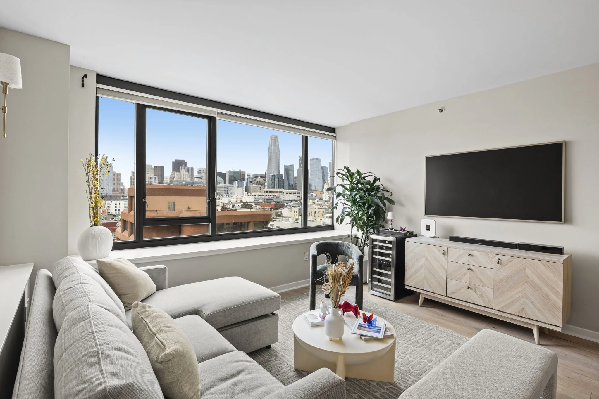 Modern living room with large windows, city skyline view, beige sofa, white coffee table with decor, black TV on a light wood cabinet, potted plant, small wine cooler, and minimalistic decor.