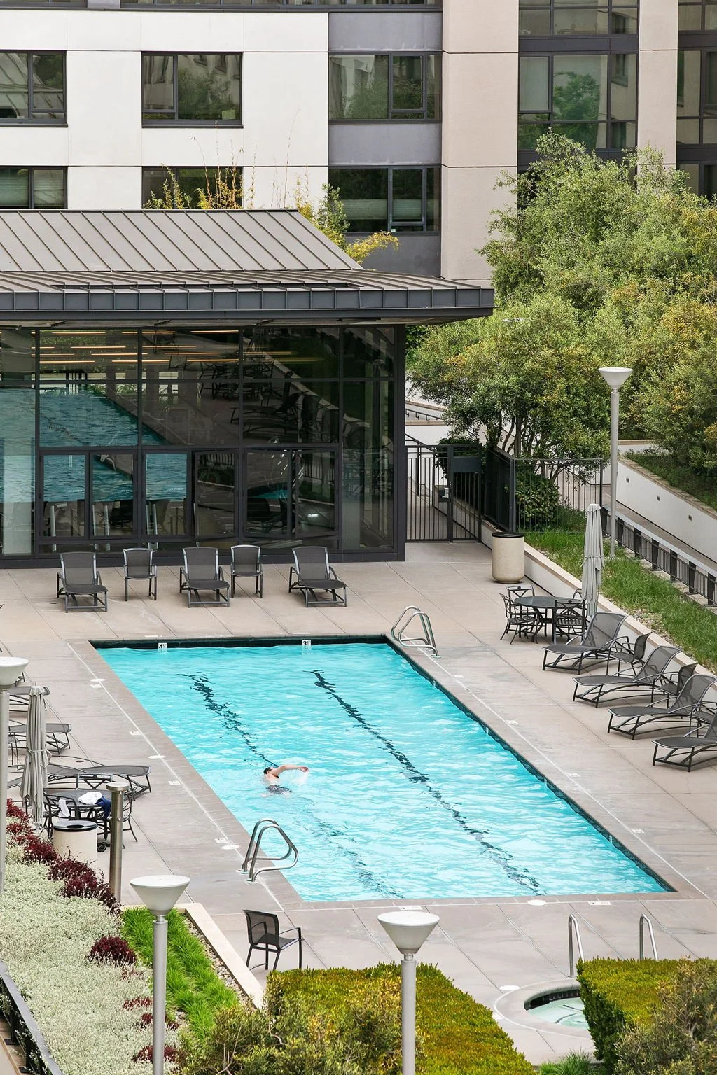 A person swimming in an outdoor pool at a modern apartment complex with lounge chairs around the pool and trees in the background.