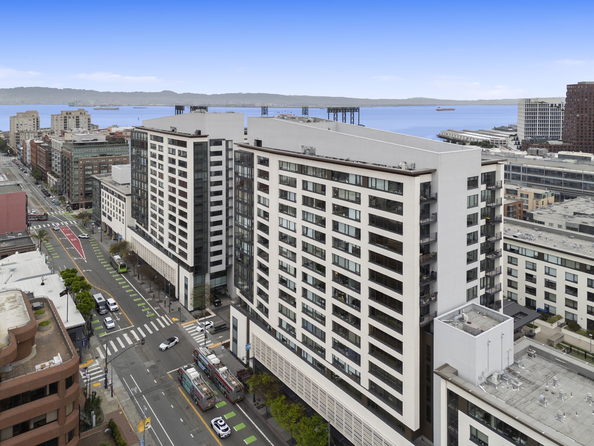 View of a city street with high-rise buildings, buses, cars, and a waterfront in the background showing water and ships under a clear blue sky.