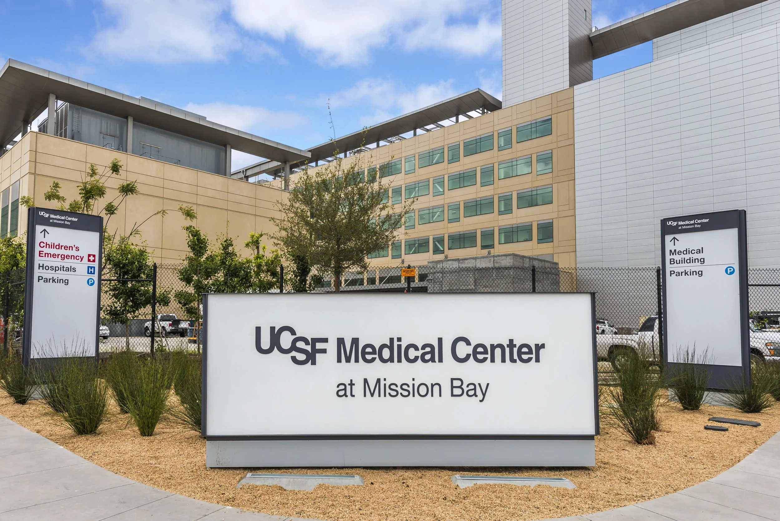 Exterior view of UCSF Medical Center at Mission Bay with signs indicating directions to children's emergency, hospitals, parking, and medical building, with modern buildings and a partly cloudy sky in the background.