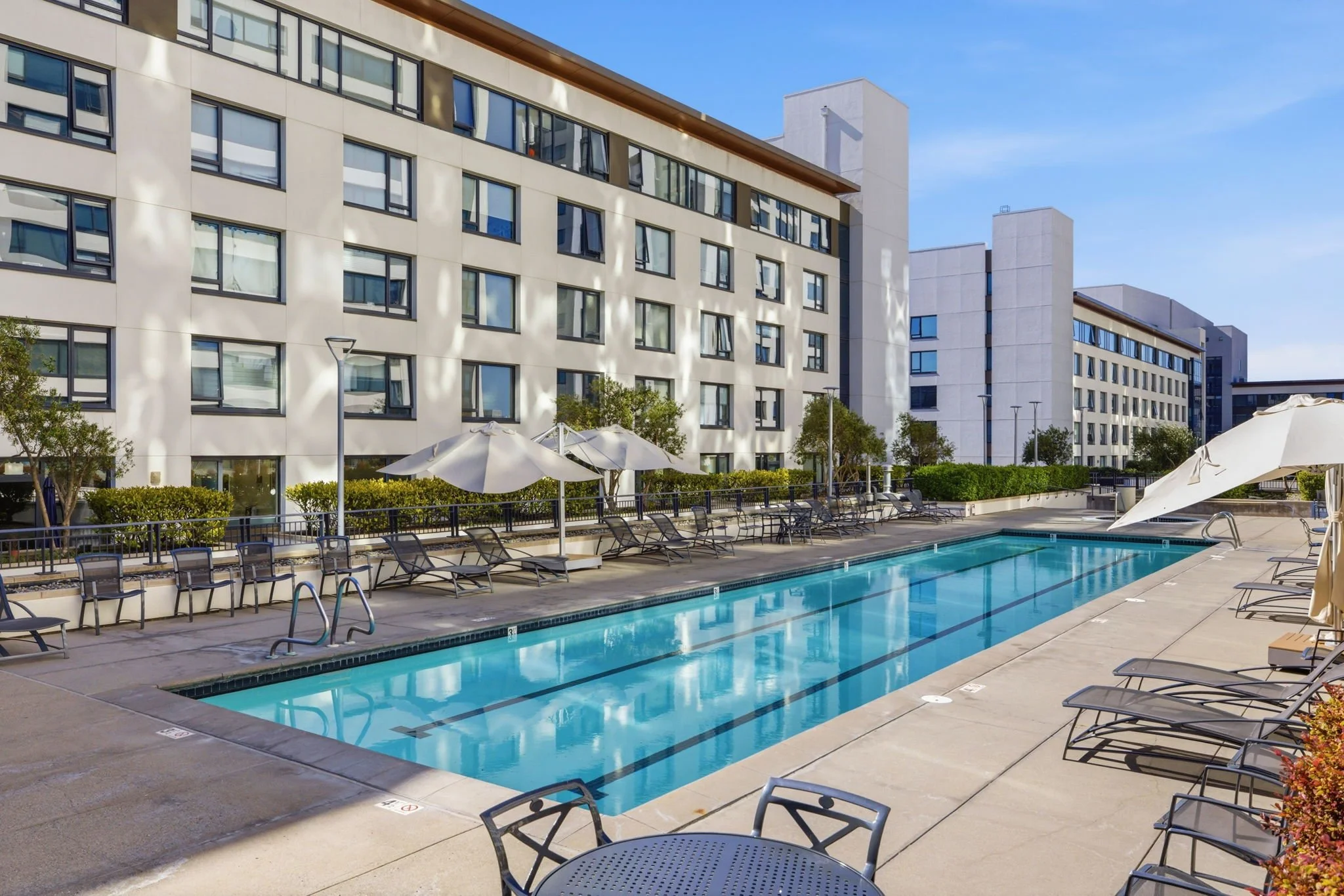 Empty outdoor swimming pool in a hotel courtyard with lounge chairs and umbrellas, surrounded by modern white hotel buildings under a clear blue sky.