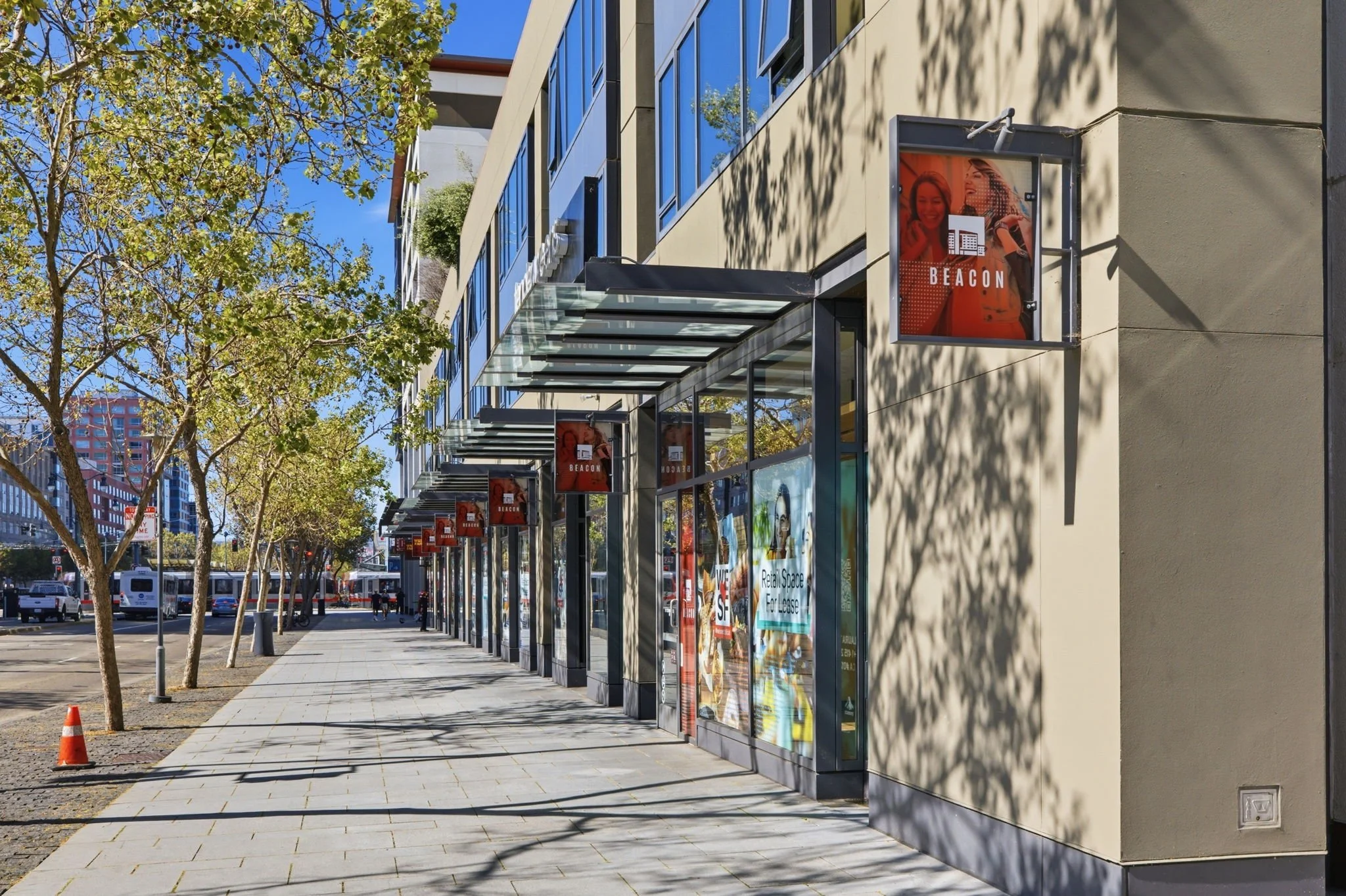 Street view of a commercial building with glass storefronts, trees shading the sidewalk, and multiple red signs with the word 'BEACON'.