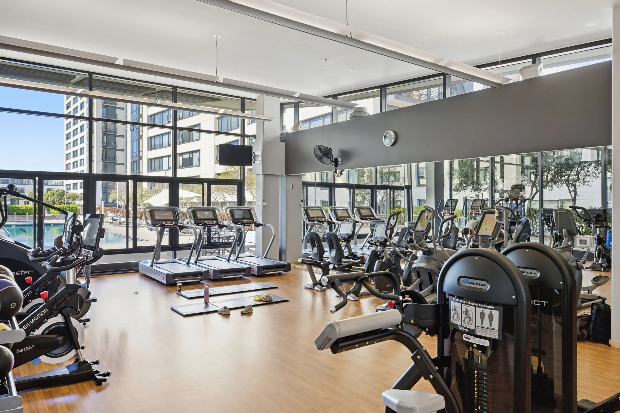 Indoor gym with large windows, treadmills, exercise bikes, and weight machines on wooden floor.