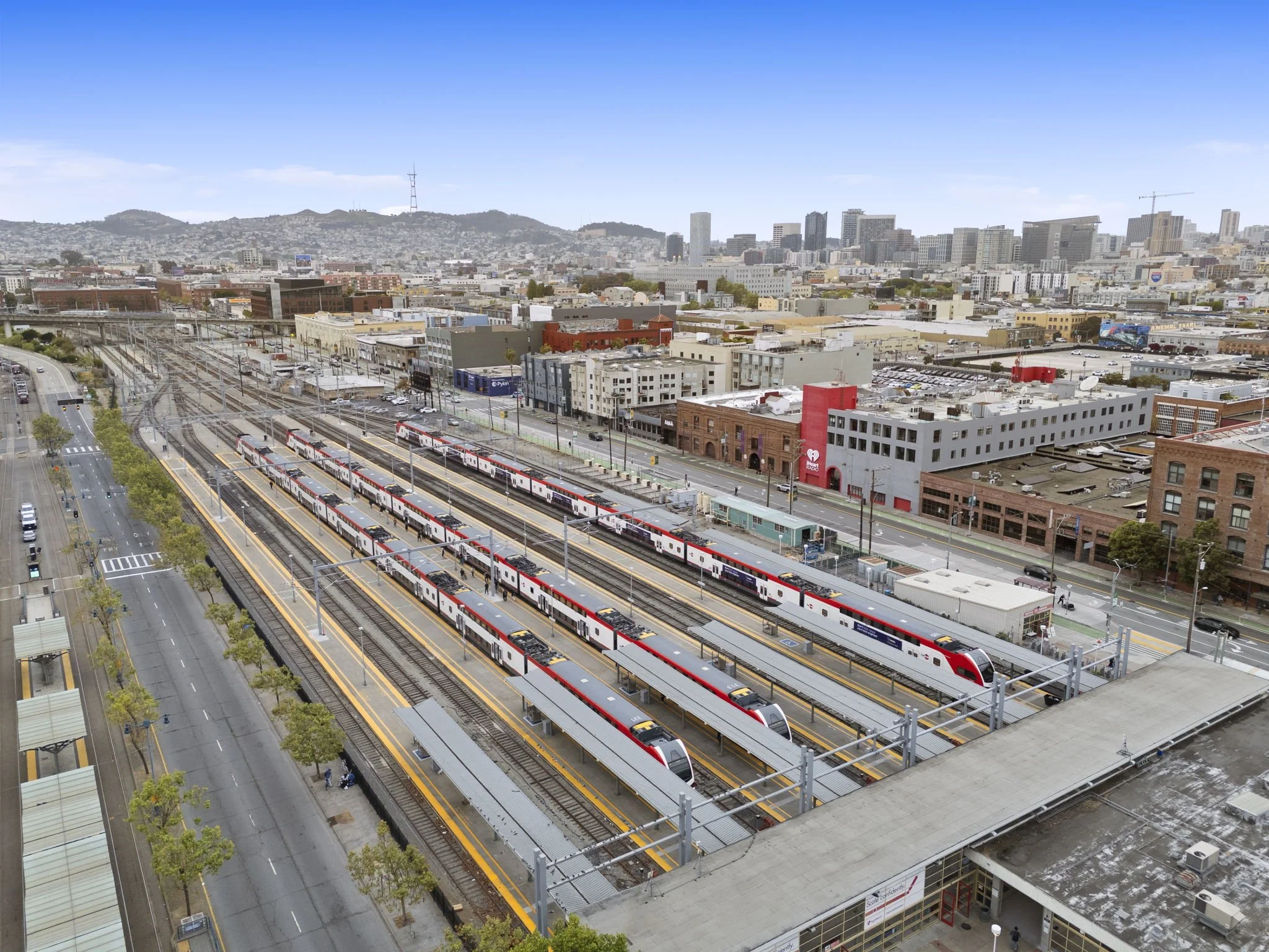 Bird's eye view of a train yard with multiple red and white trains on tracks, adjacent to a cityscape with buildings and hills in the background.