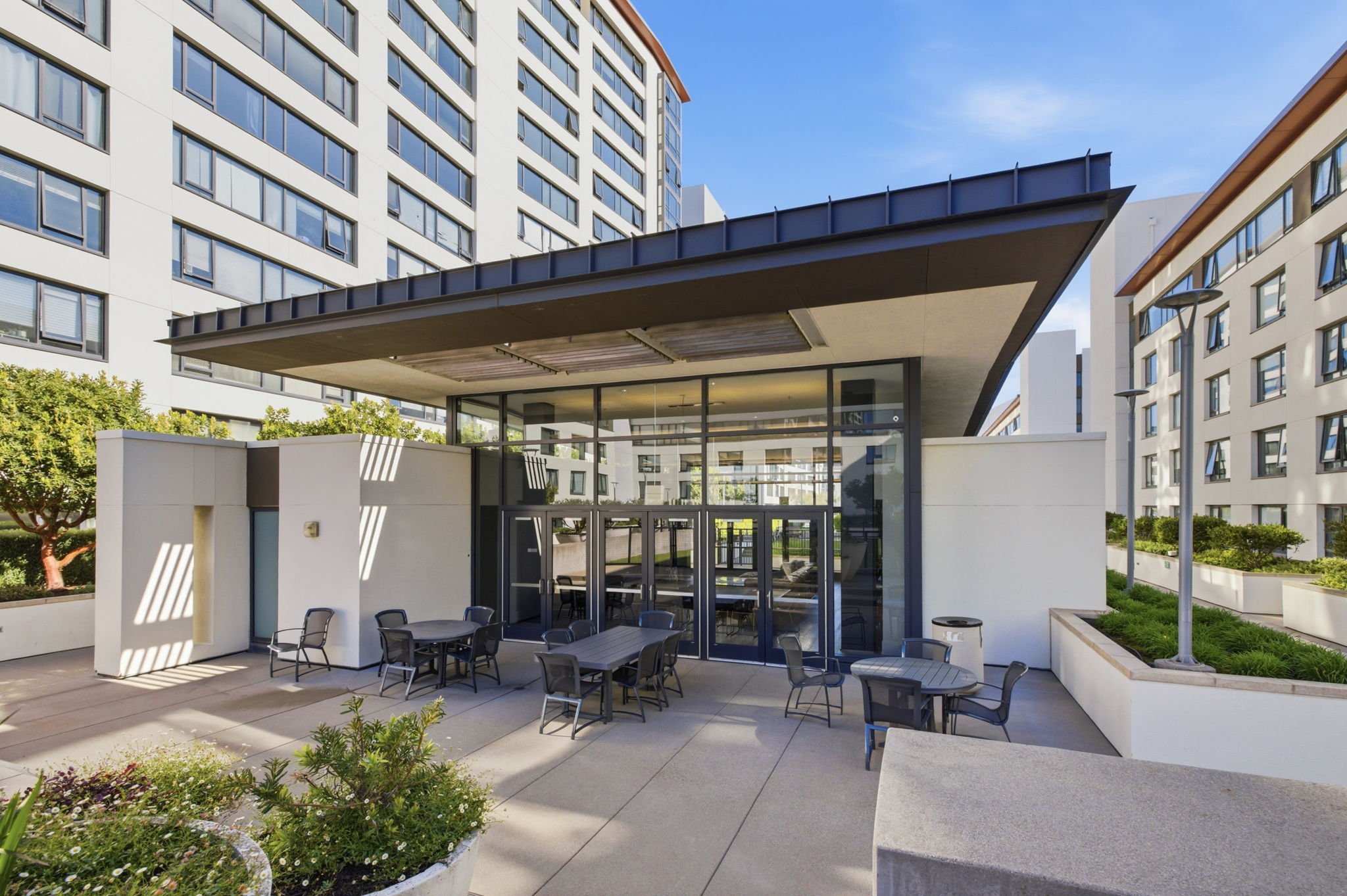 Outdoor seating area with tables and chairs in front of a modern glass entrance to a high-rise building complex, with clear blue sky overhead.