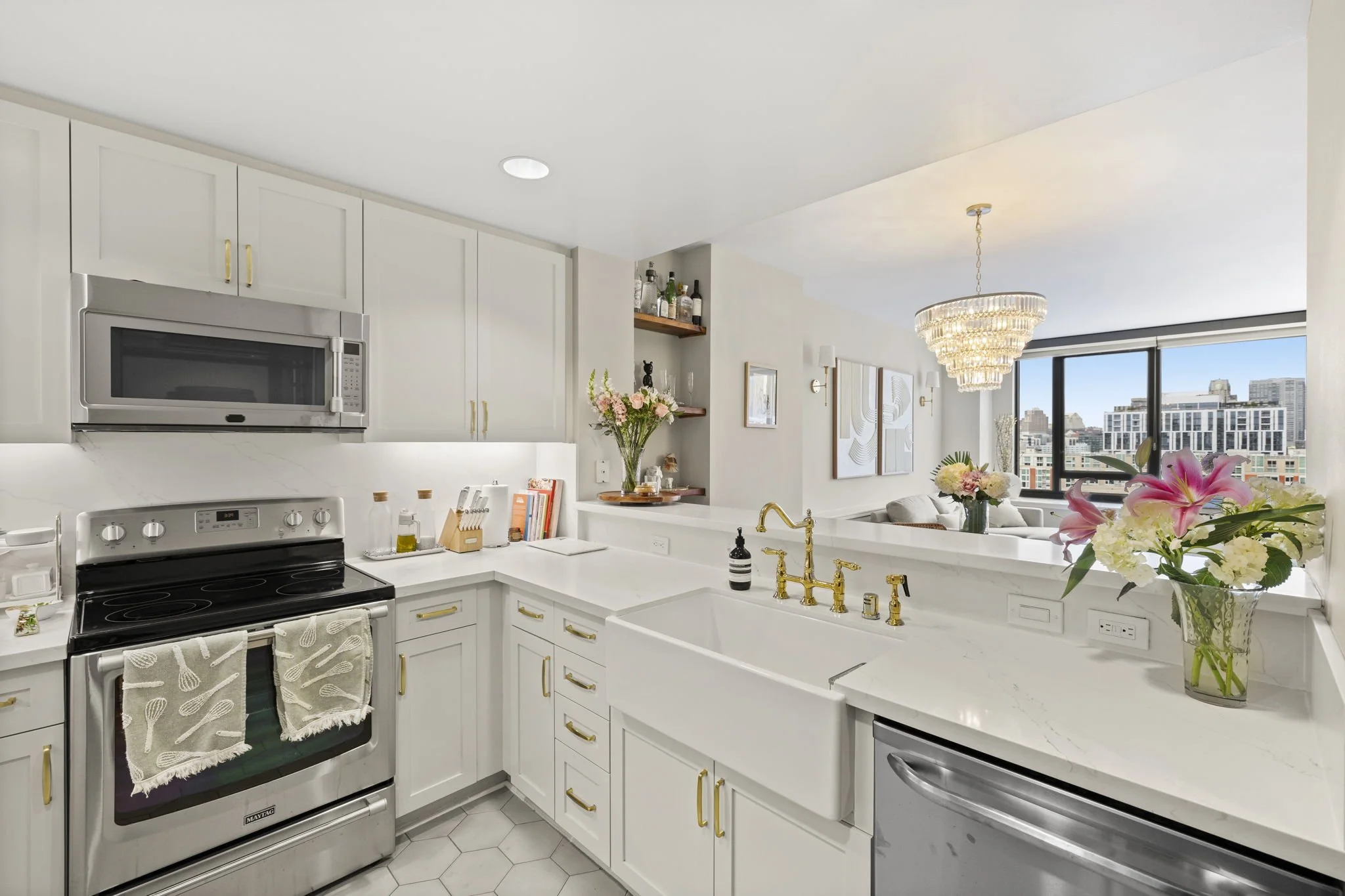 Modern kitchen with white cabinets, marble countertops, and gold hardware, overlooking a living room with large windows and a city view, decorated with flowers and a chandelier.