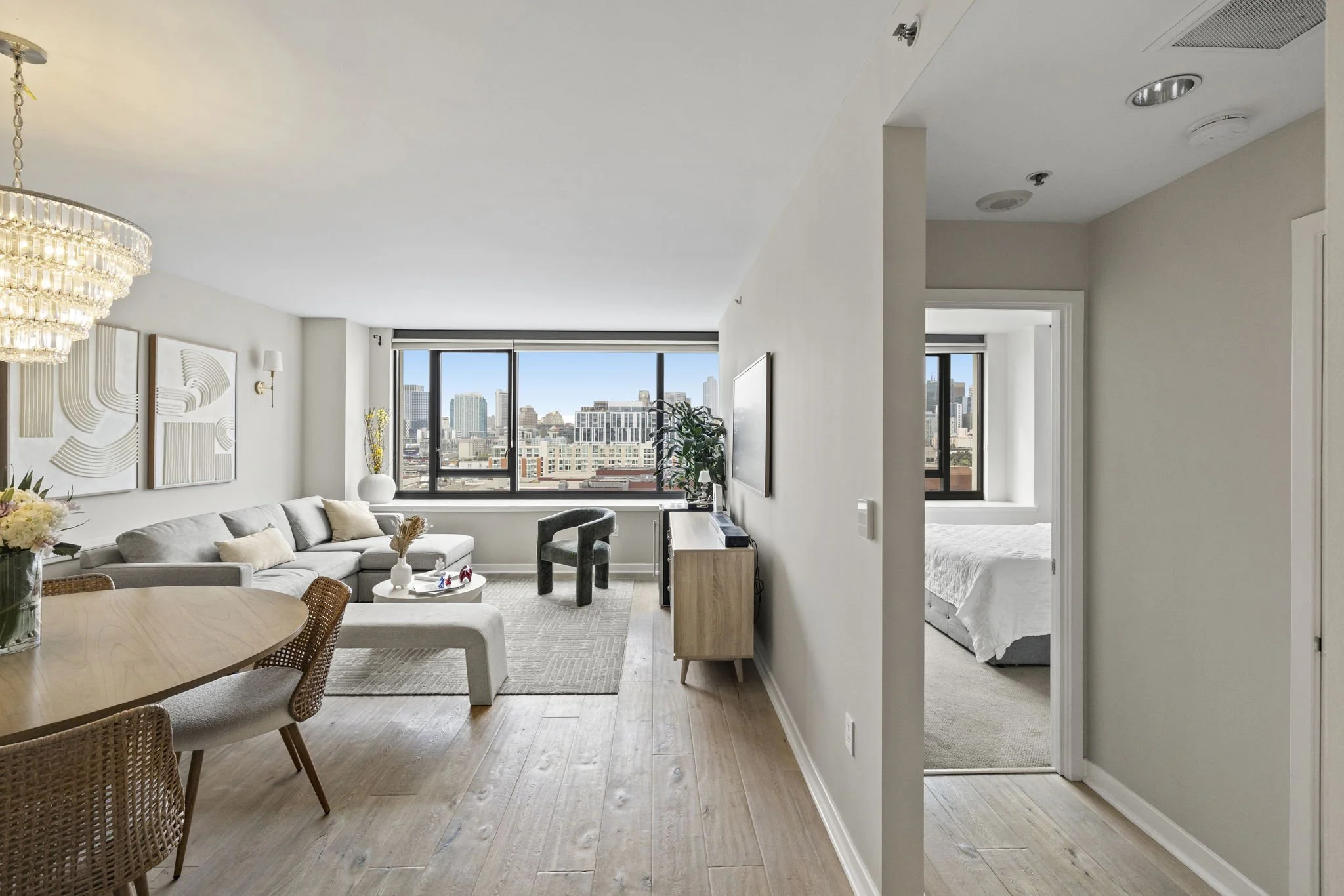 Modern apartment living room with large windows, gray sofa, dining table, decorative wall art, and a view of city buildings.