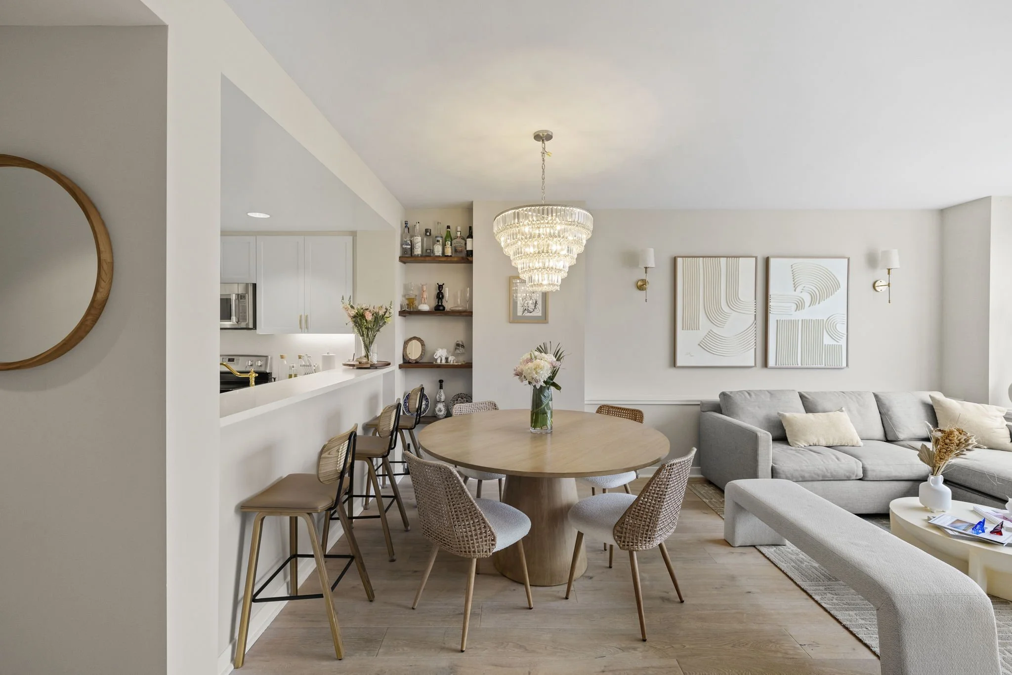 Open concept living and dining area with a round wood dining table, six chairs, a chandelier, a grey sectional sofa, artwork on the wall, and a view into the kitchen with white cabinets and bar stools.