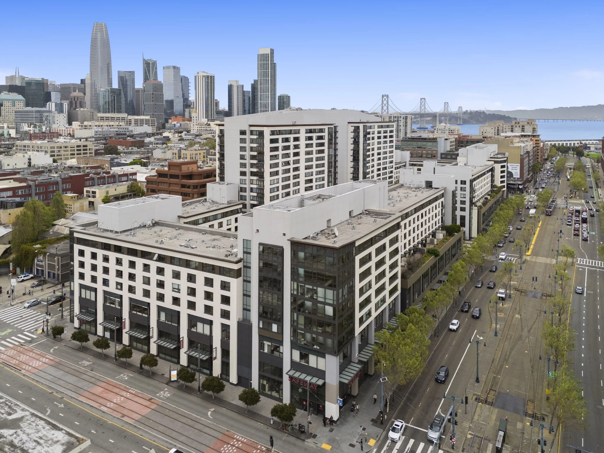 Aerial view of a cityscape featuring modern mid-rise and high-rise buildings, with a bridge spanning across a body of water in the background, and a wide street lined with trees and parked cars.