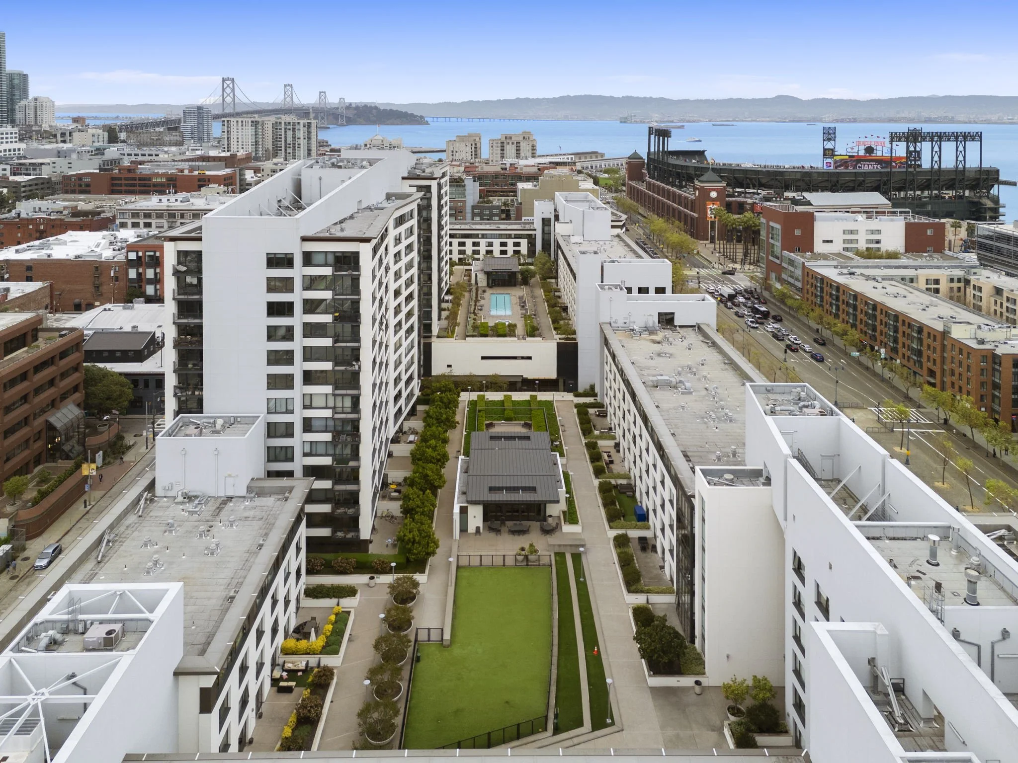 A high-angle view of a cityscape featuring modern white residential buildings, a central courtyard with green lawn, trees, and a swimming pool, with the San Francisco Bay and Bay Bridge in the background.