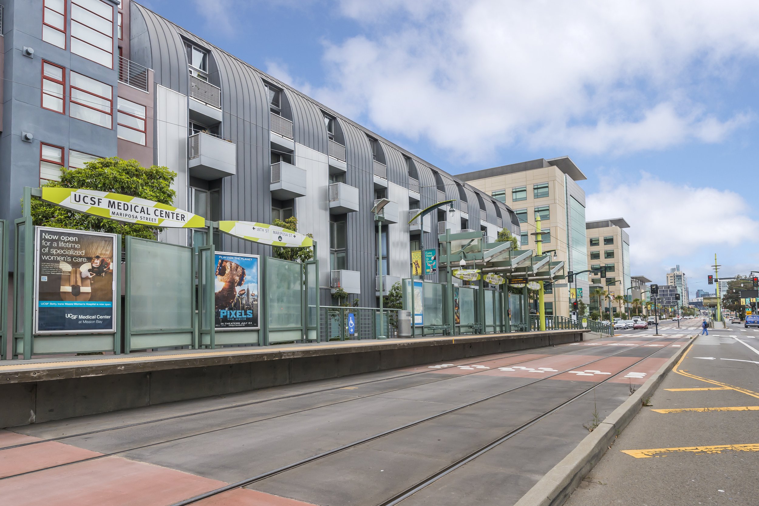Urban street scene with a modern UCSF Medical Center building, tram tracks, bus stop, and city traffic against a partly cloudy sky.