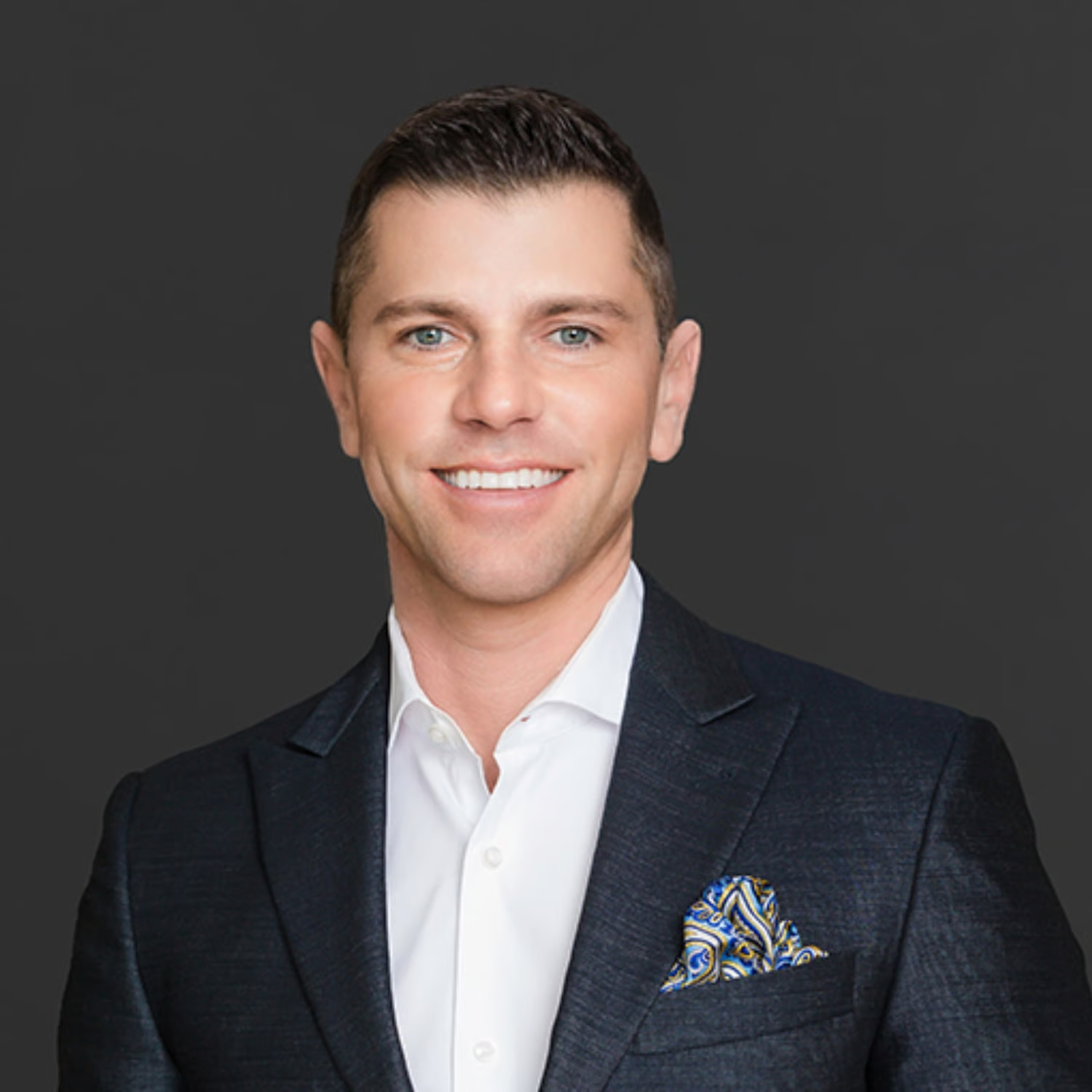 Headshot of a man in a dark suit smiling against a dark gray background.