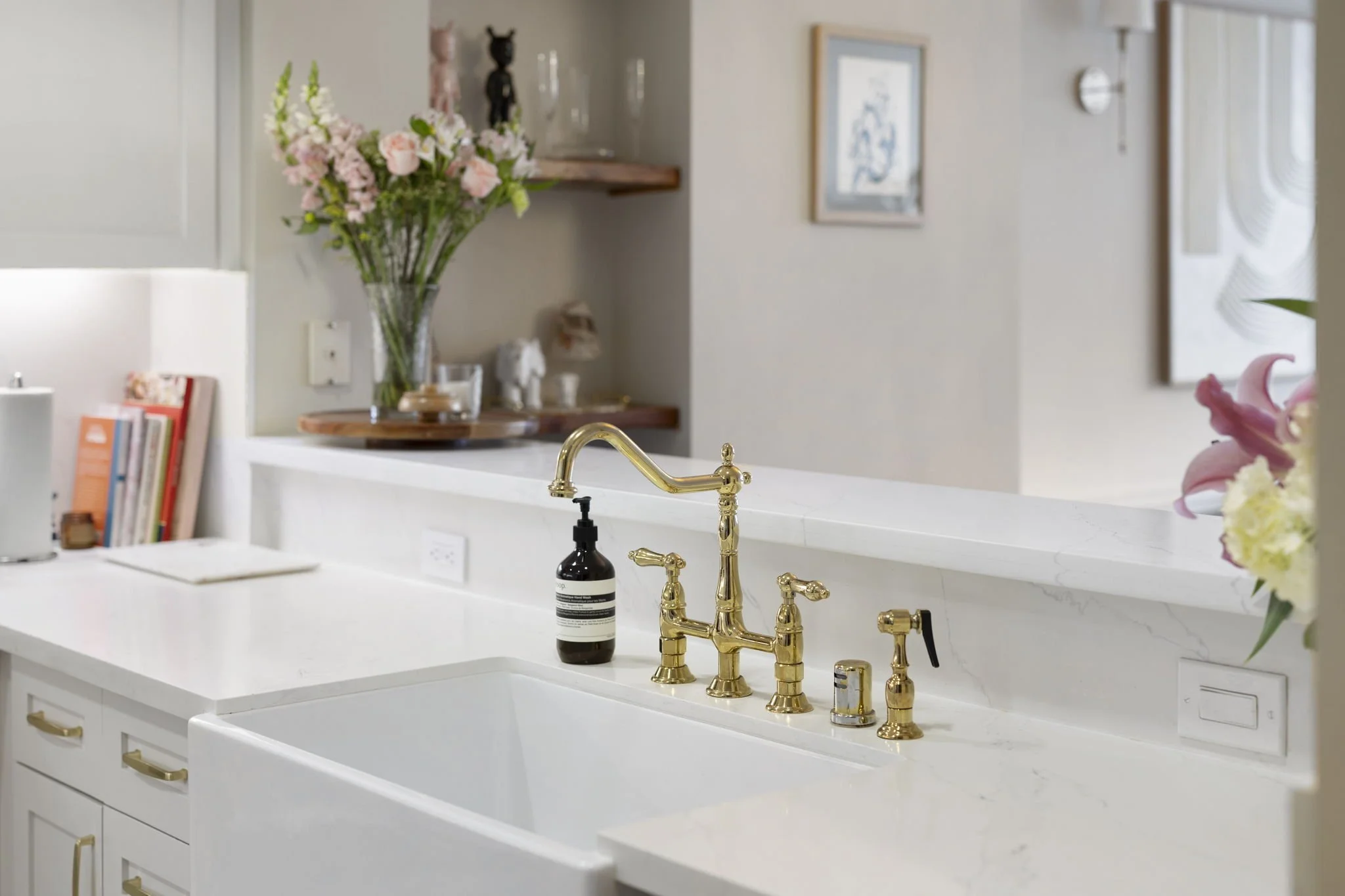Kitchen sink with gold faucet, soap dispenser, and a white farmhouse-style sink, with pink and white flowers in the foreground and decorative items on shelves in the background.
