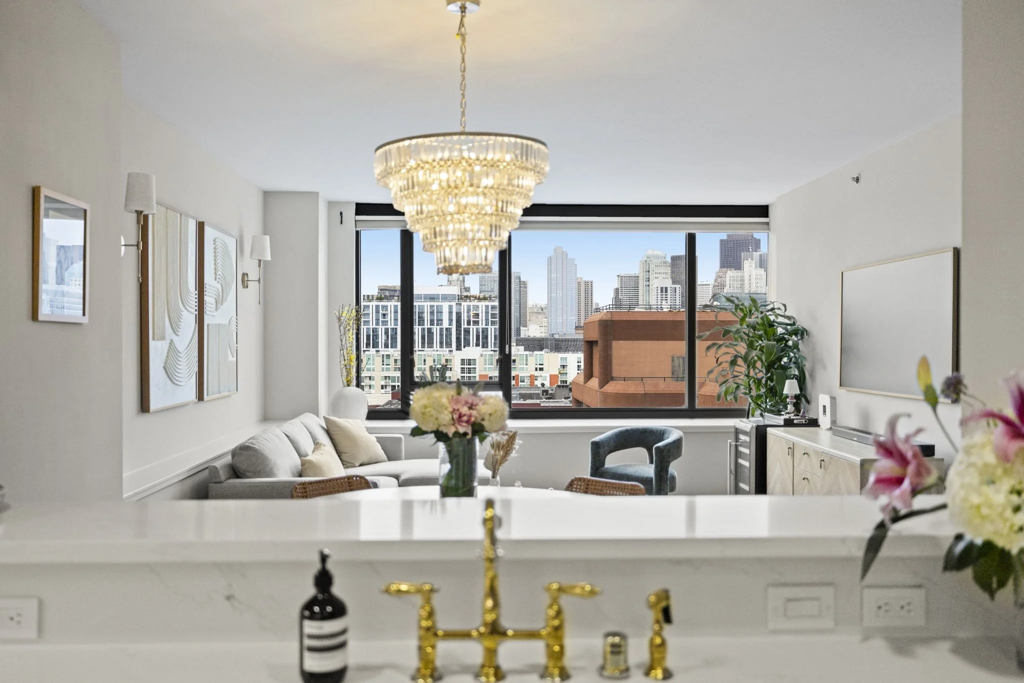 Modern living room with white walls, gold chandelier, artwork, gray sofa with pillows, large windows showing city skyline, black armchair, plants, and a sideboard.