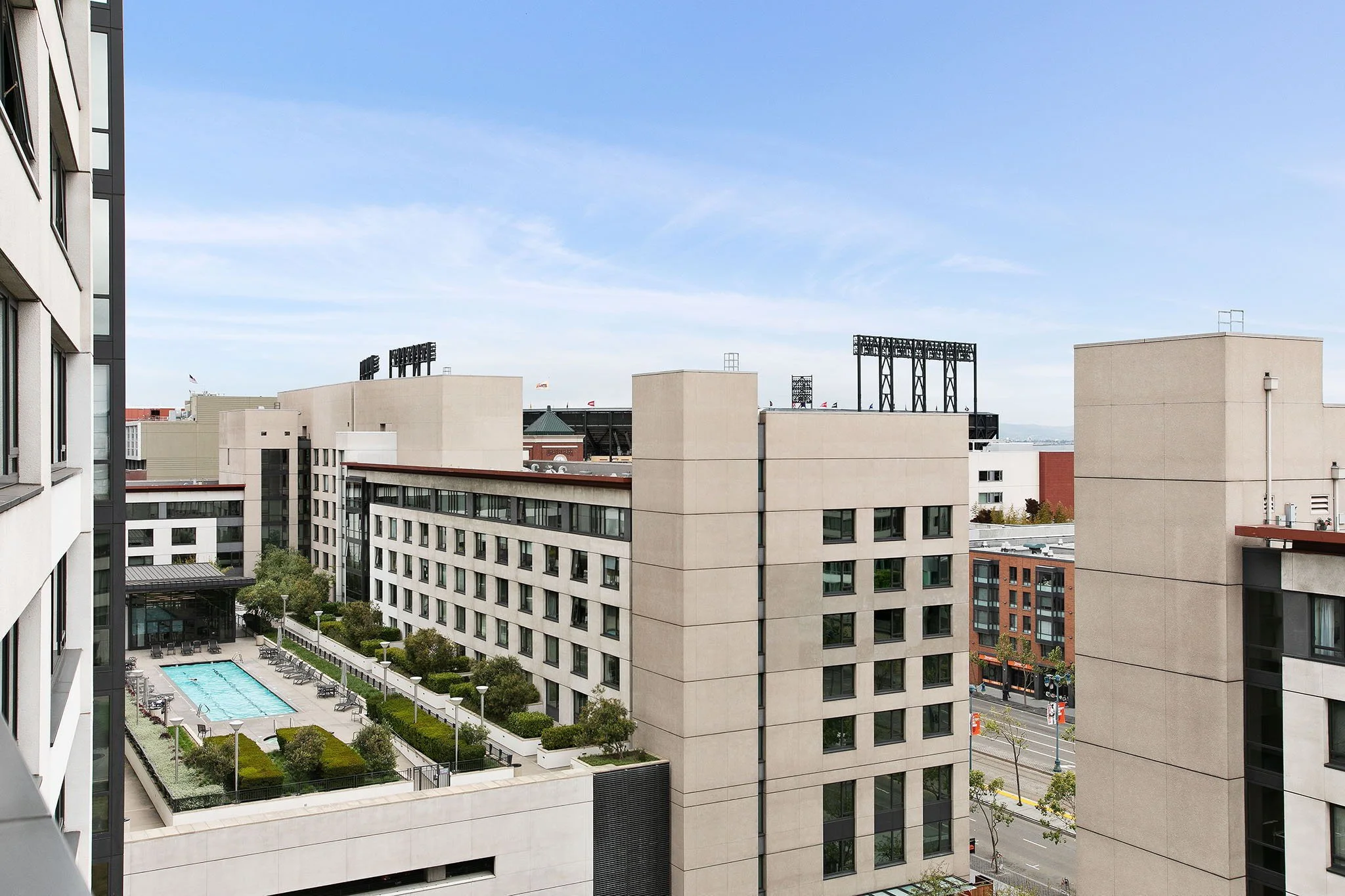 View of a modern apartment complex with a central pool area, surrounded by trees and shrubbery, with other buildings and a street visible in the background under a blue sky.