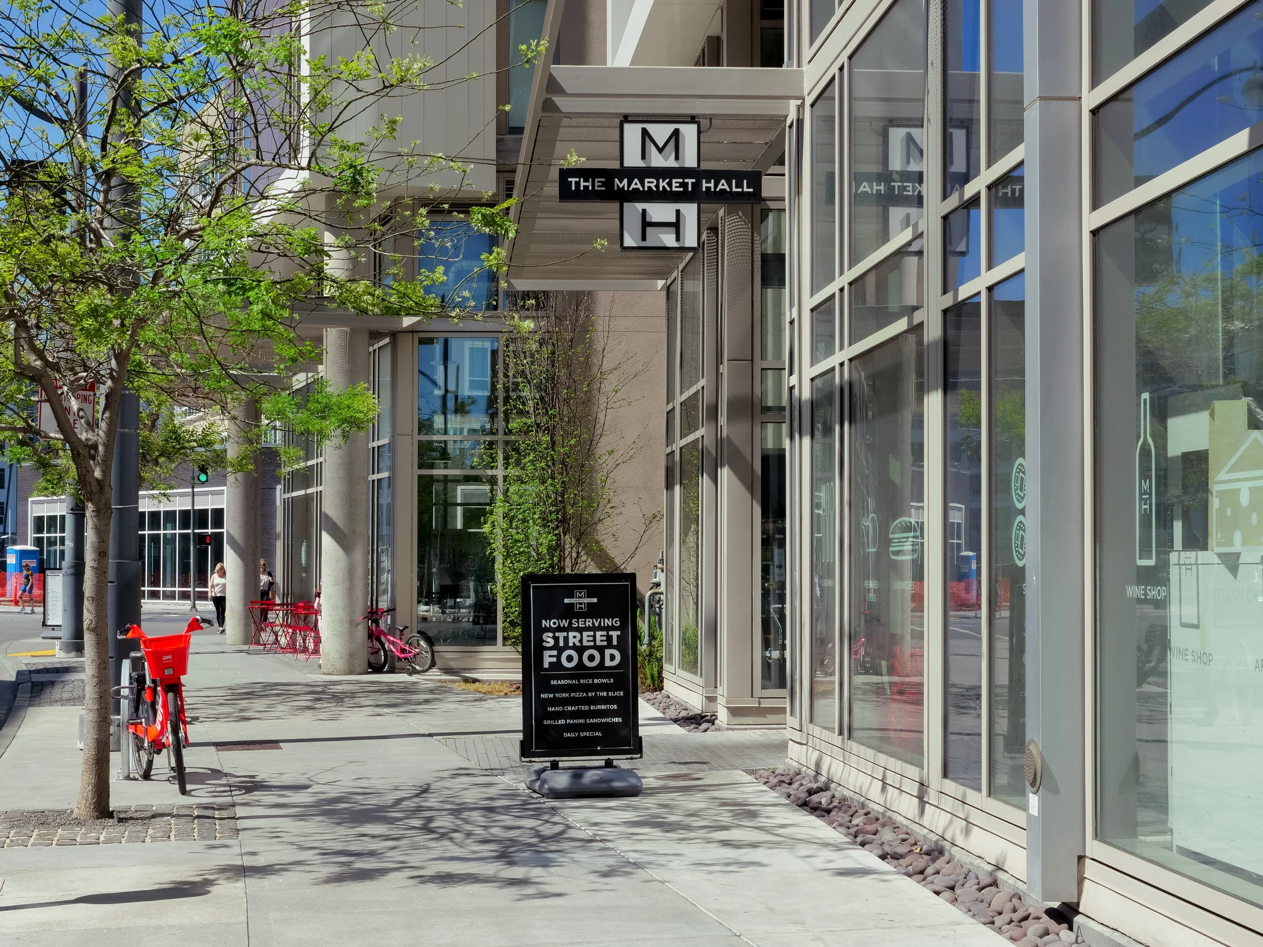 Sidewalk outside a modern building with a sign advertising street food, bicycles parked, and a tree with green leaves.