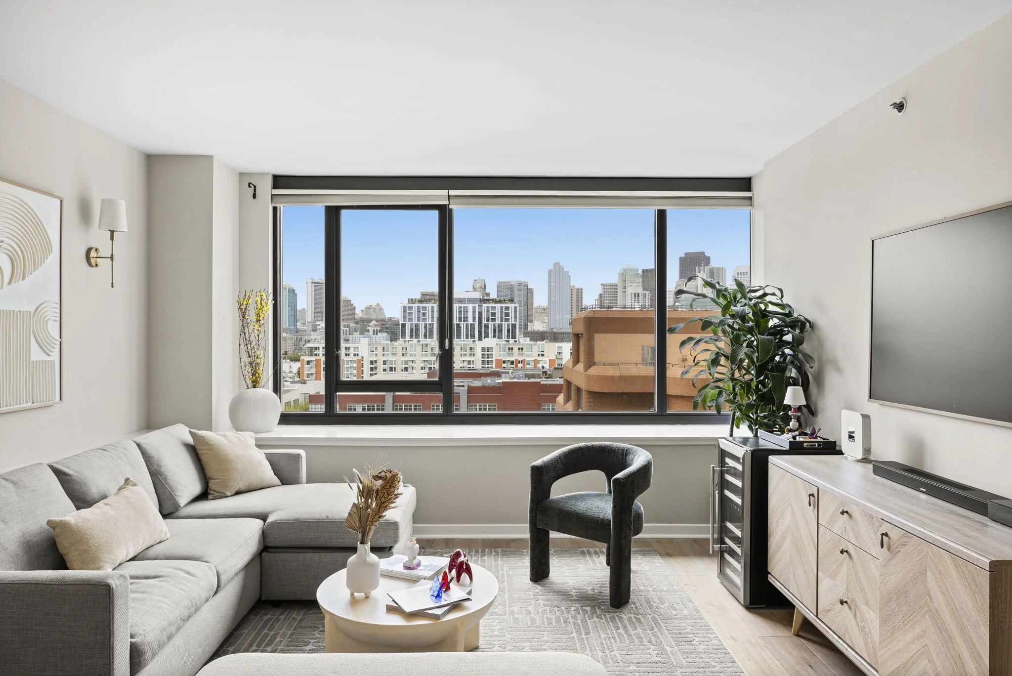 Modern living room with gray sofa, black armchair, white coffee table, large window with city skyline view, decorative plants, wall art, and wooden sideboard.
