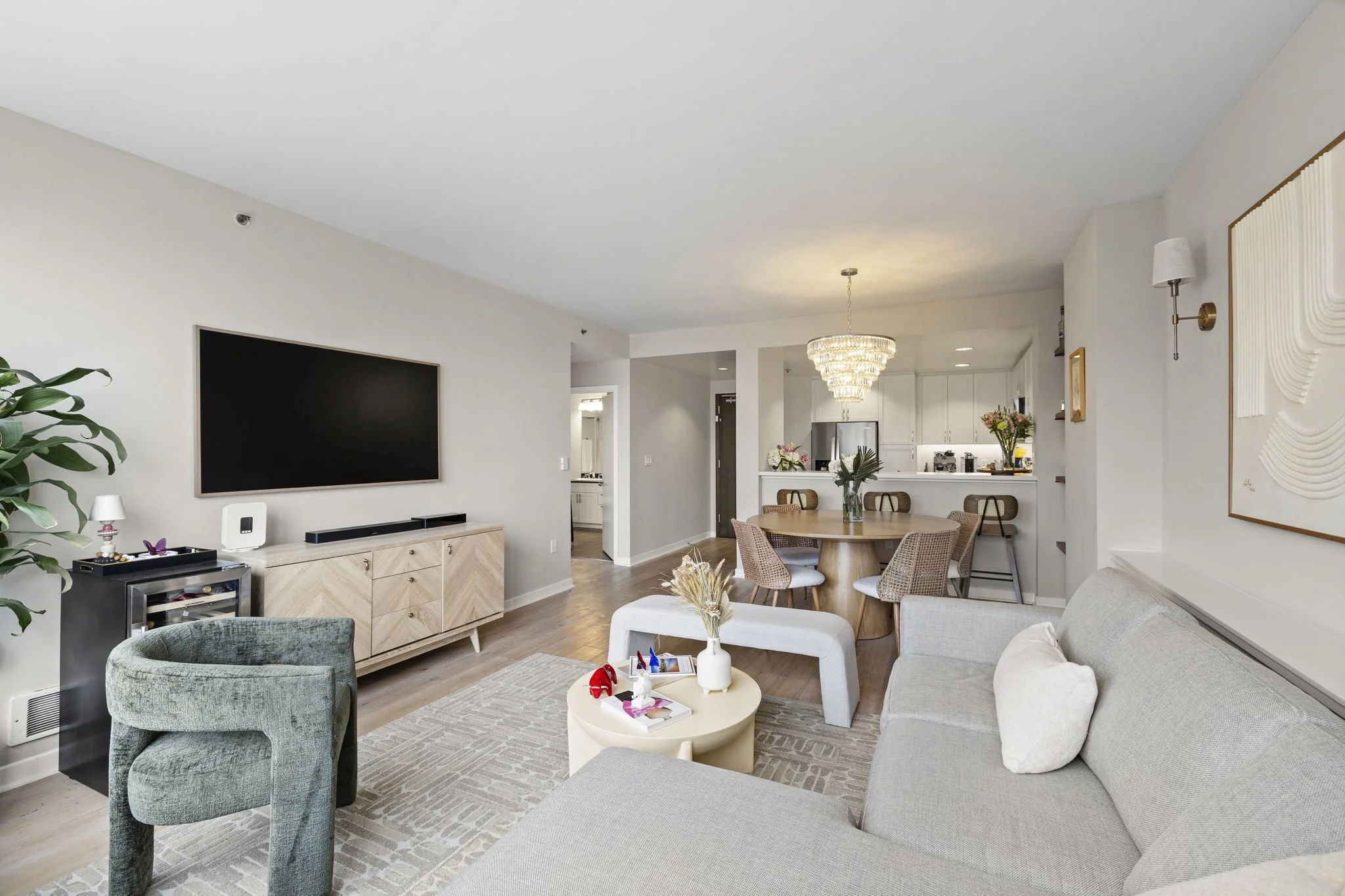 Modern living room with a large TV on a wooden console, gray sofa, green accent chair, round coffee table, and a dining area with a chandelier, wooden dining table, and chairs.