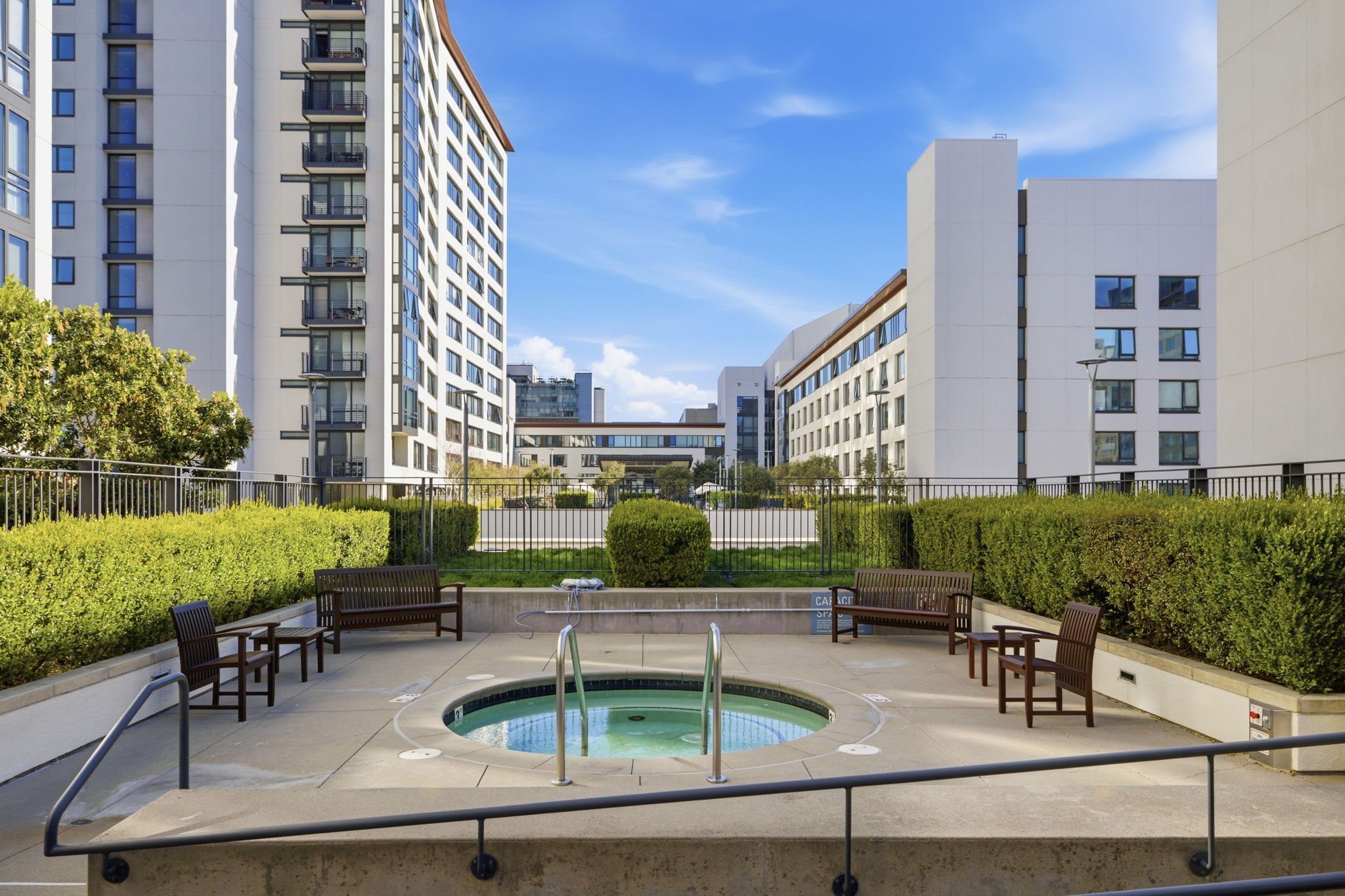 Outdoor hot tub with metal handrails, surrounded by concrete, with four wooden benches and green bushes in a courtyard of a modern apartment complex under a blue sky.