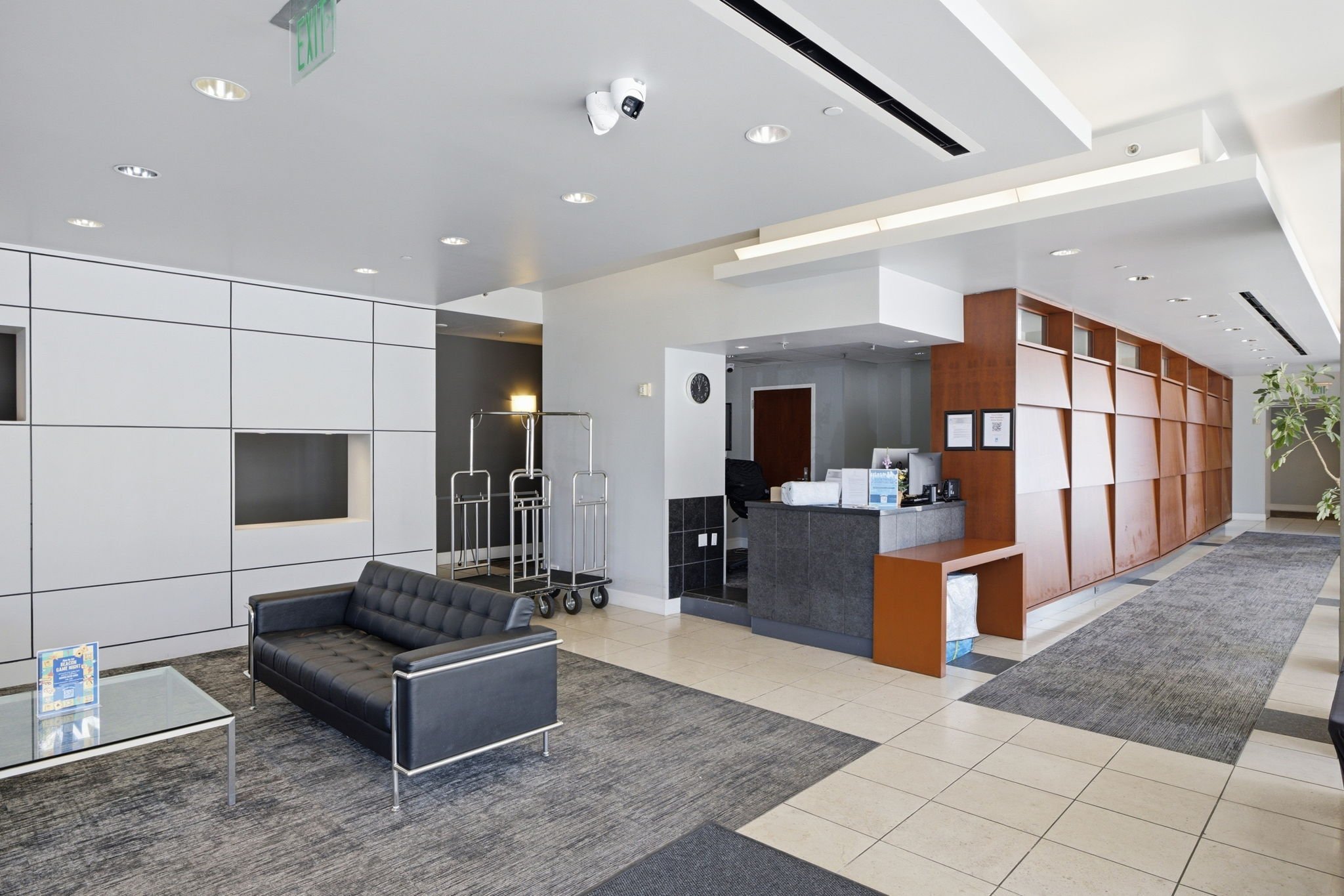 Modern hotel lobby with black leather sofa, glass coffee table, reception desk, luggage carts, wooden wall panels, and indoor plants.