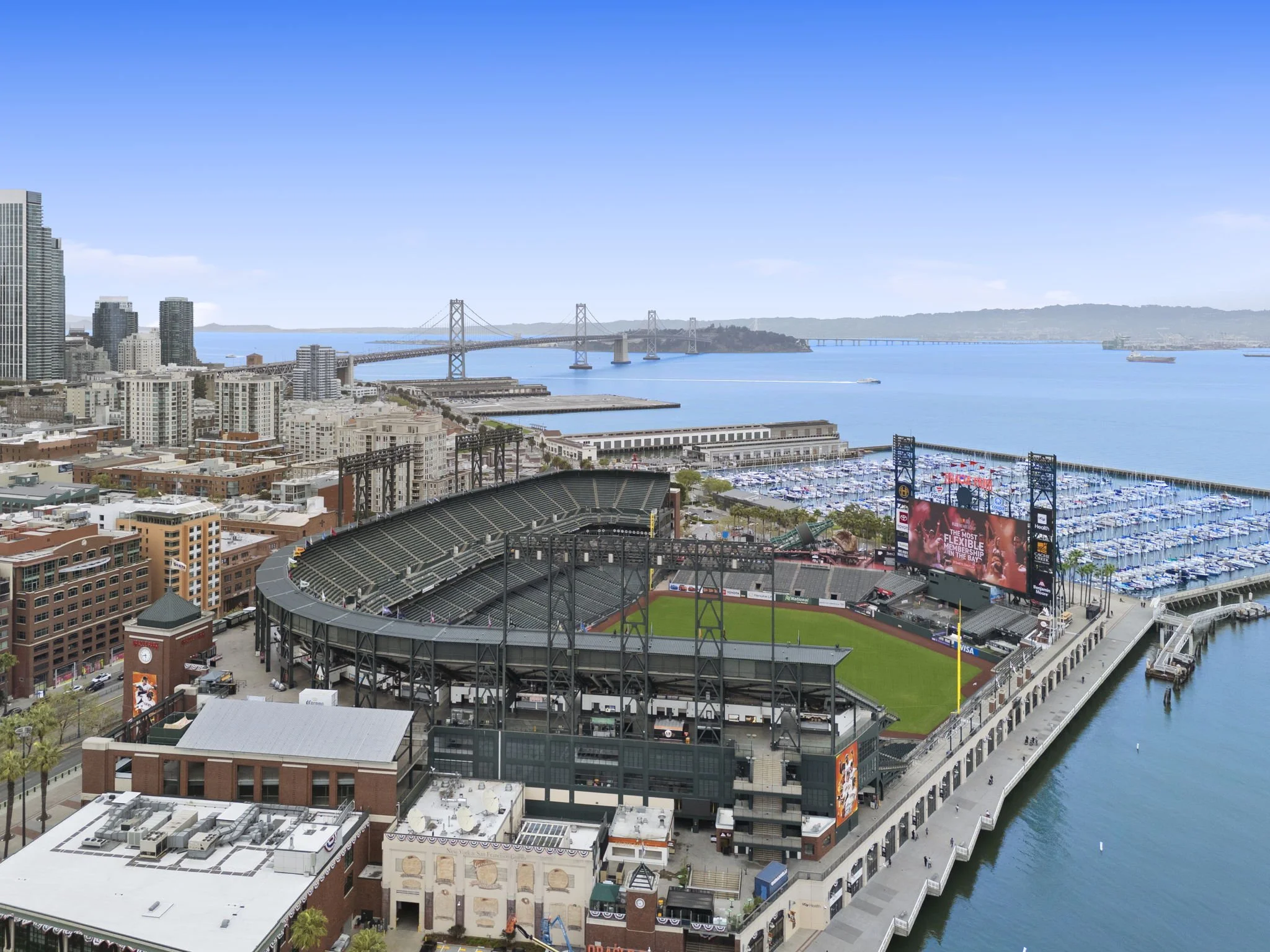 Aerial view of a baseball stadium near the water with boats in a marina, a bridge in the background, and city buildings around.