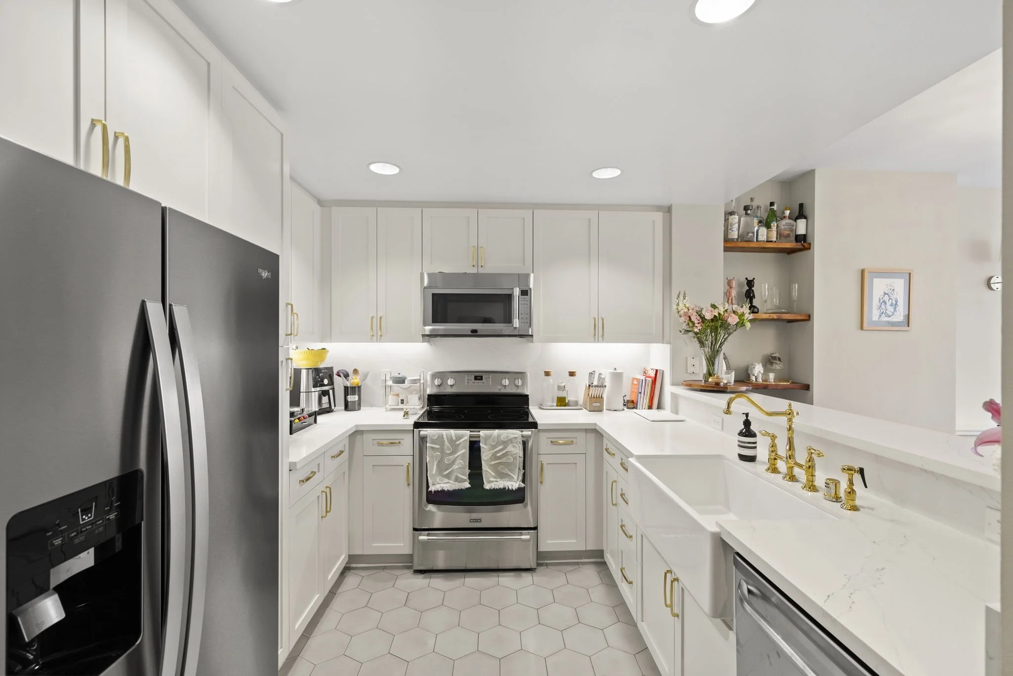 A modern kitchen with white cabinets, a stainless steel refrigerator, stove, microwave, and gold hardware. There are open shelves with bottles and decorative items, a white sink with gold fixtures, and pink flowers on the counter.