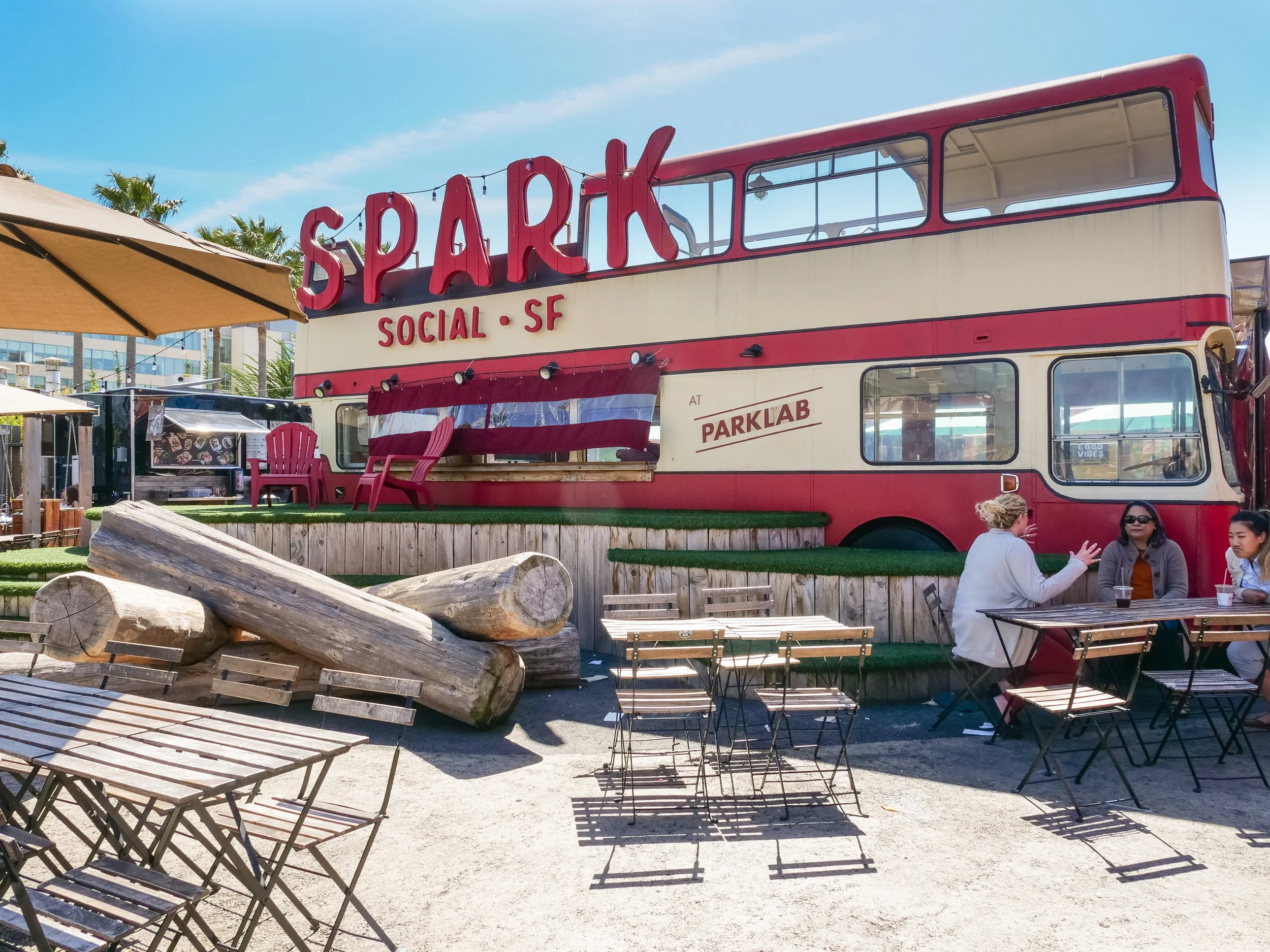 Outdoor seating area with tables and chairs, large red and beige double-decker bus with sign reading 'SPARK' and 'SOCIAL SF', people sitting and talking at a table, sunny day with blue sky, trees and buildings in background.